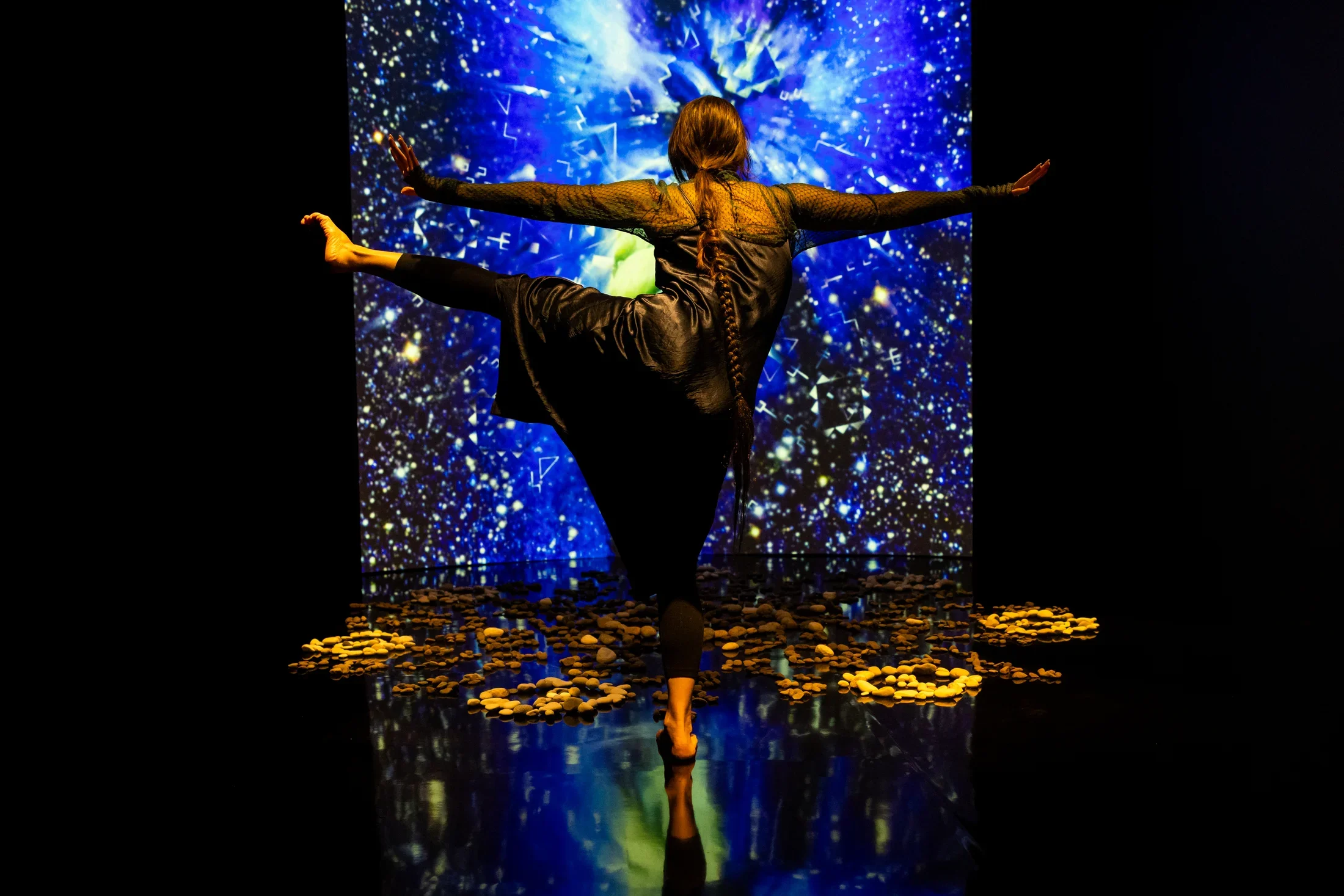 Still of a performance showcasing a woman spotlighted on stage with her arms out to both sides and her left leg raised to hip height with foot pointed. A brightly colored geometric pattern in the background is juxtaposed by a black wall.