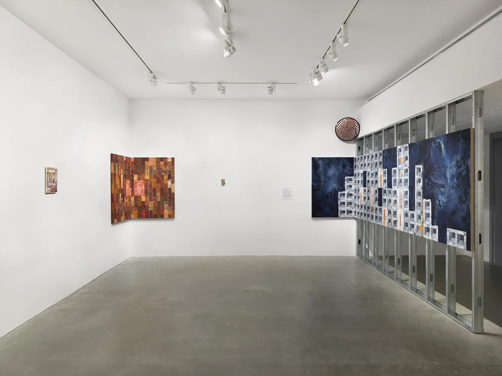 A blue cinder block-patterned painting wraps from scaffolding to the wall; a painted mirror and three other paintings hang throughout the gallery.