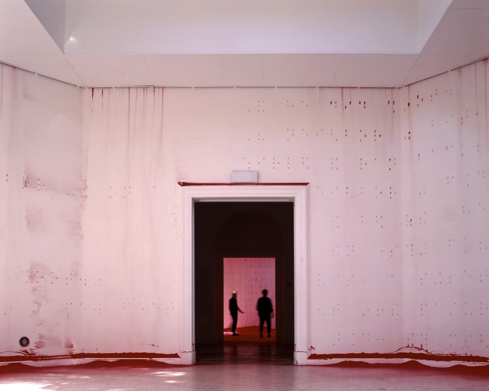 An open white-walled room with braille raised bumps across the walls. The silhouettes of two people can be seen through the dark doorway in the center of the image.