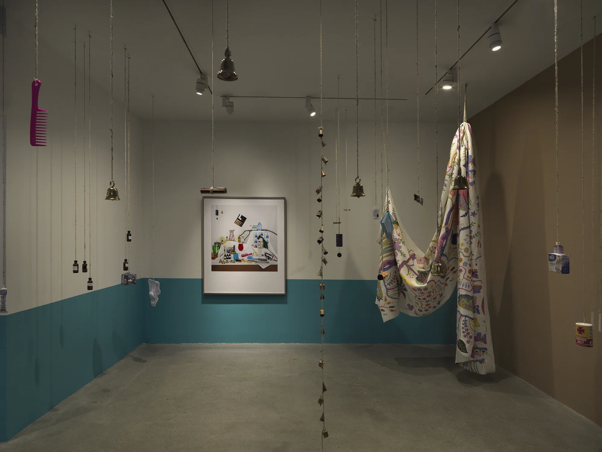 A dimly lit white walled gallery with the bottom half of the wall painted turquoise. A collection of everyday items hanging from strings throughout the space and a large black framed color photograph hangs on the back wall. A large printed sheet of fabric hangs on the right side of the frame. 