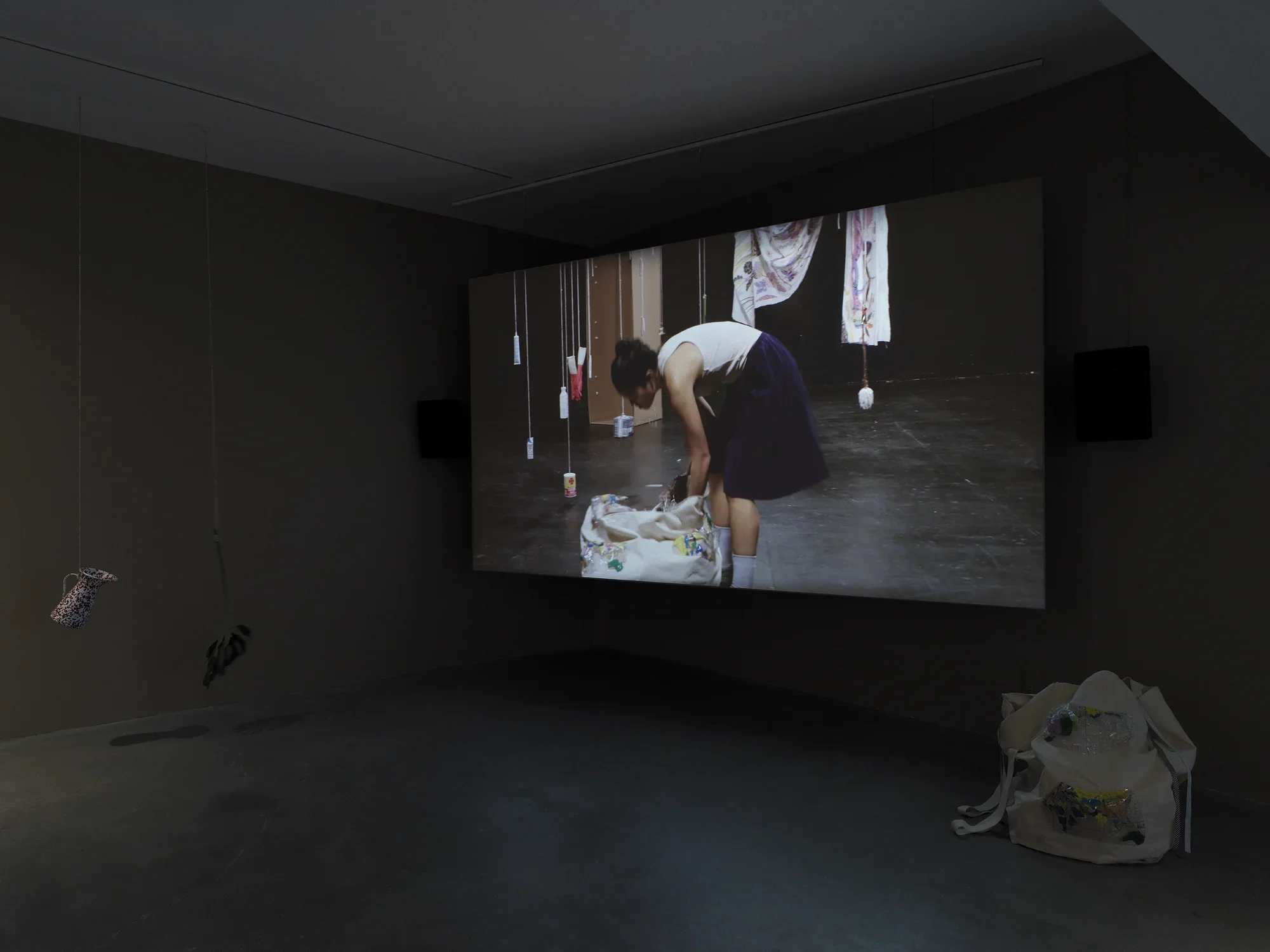 Dark open gallery with a video projected on a large standing screen in the back of the space. There is a collection of everyday items hanging from strings throughout the gallery. A tan canvas bag sits on the floor to the right of the screen.