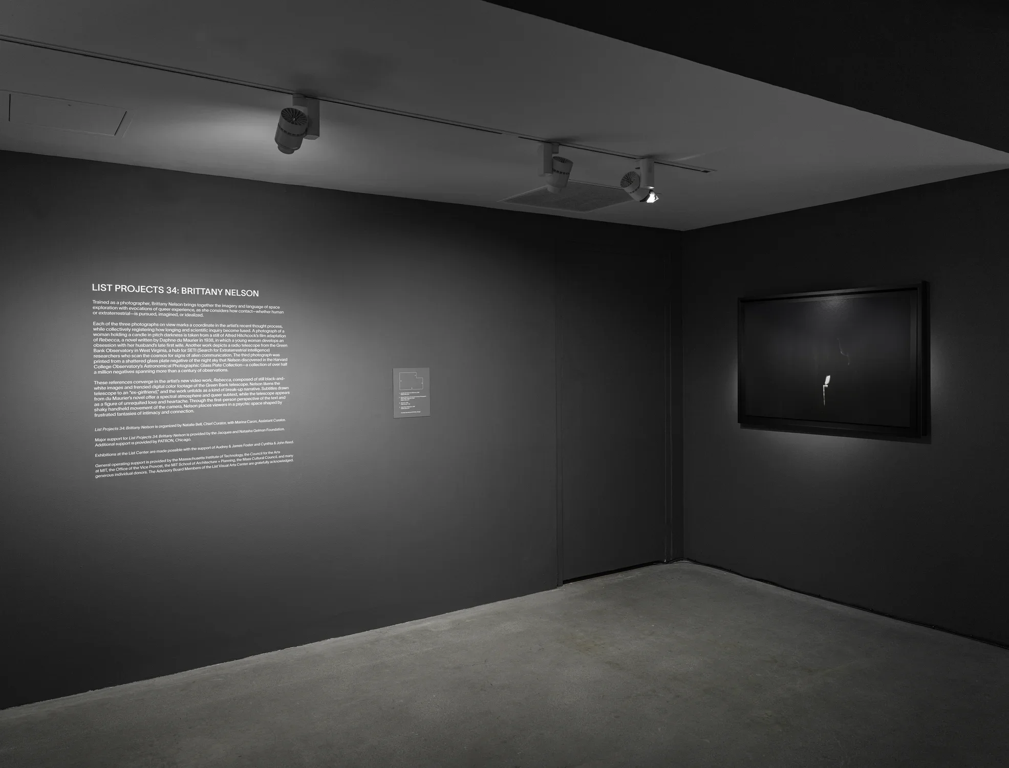 A dimly lit gallery room with dark gray walls features a text panel on the left wall. A framed artwork with a subtle light is on the right wall.