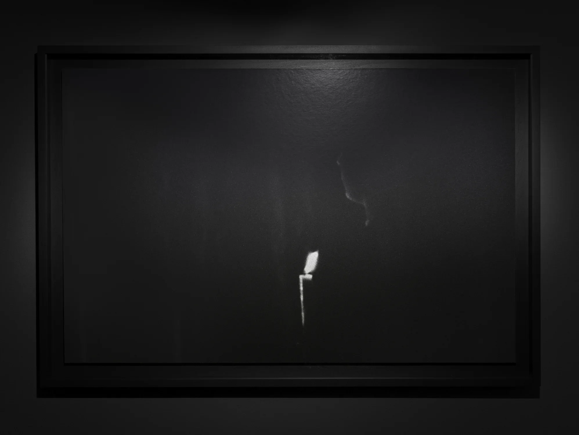 A black-and-white photo of a figure holding a candle in complete darkness. The image is predominantly black, with a lit candle at the bottom center of the frame. A faint silhouette of the figure is partially illuminated by the candlelight.