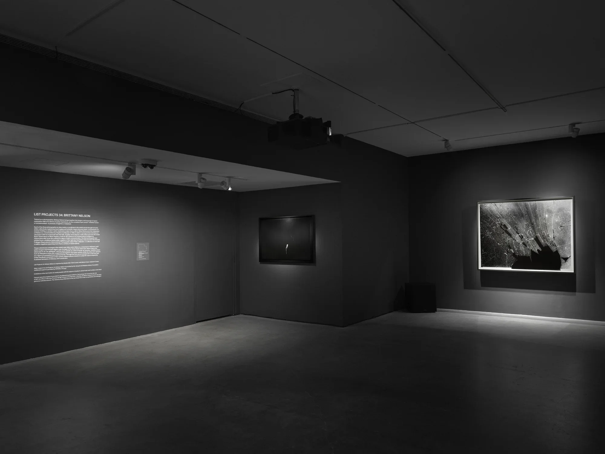 A dimly lit room with dark walls featuring two artworks. The left wall shows text in white. The right wall displays a large, abstract, black-and-white photograph.