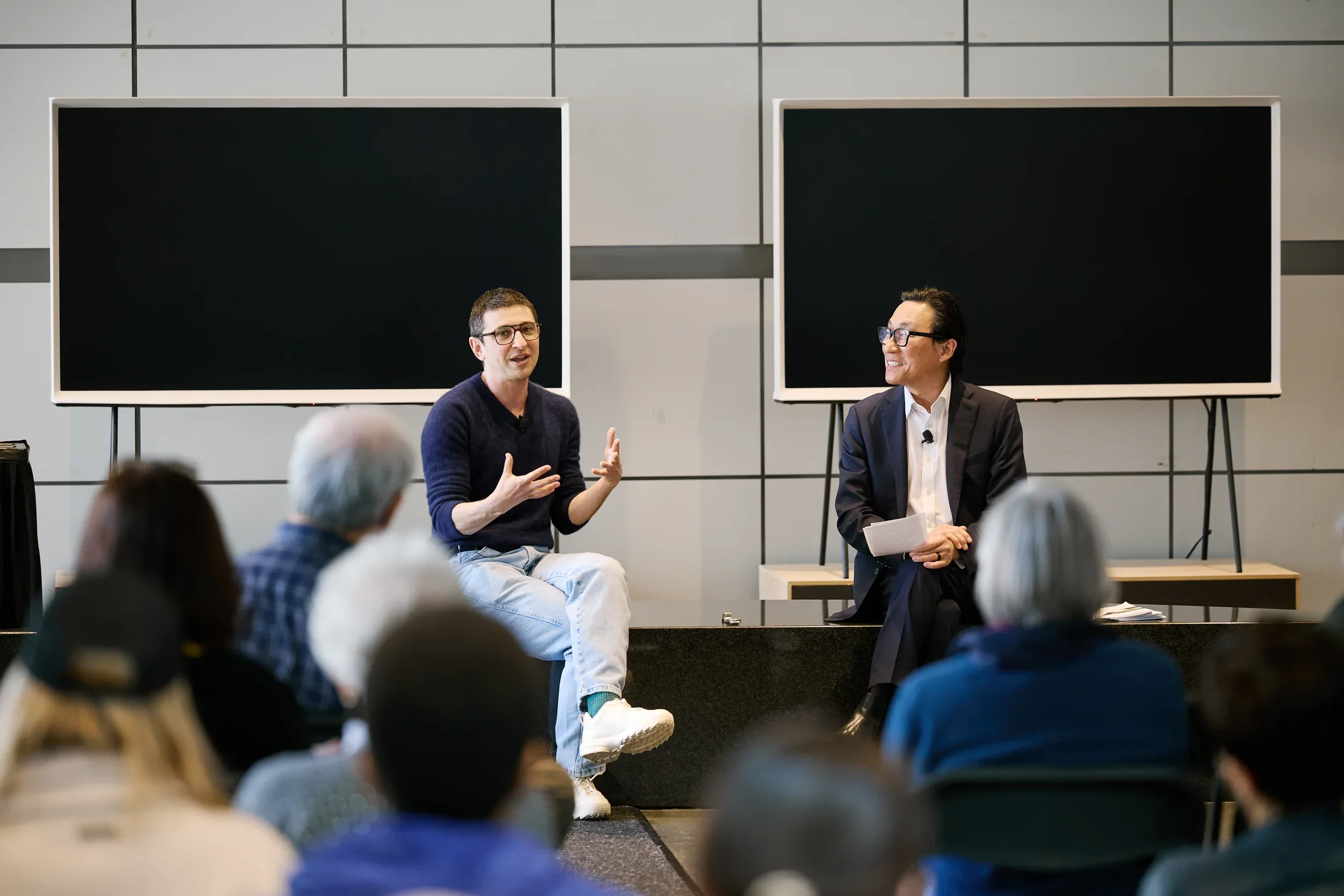 Two people sit on a bench in front of two screens speaking to an audience sitting facing them.