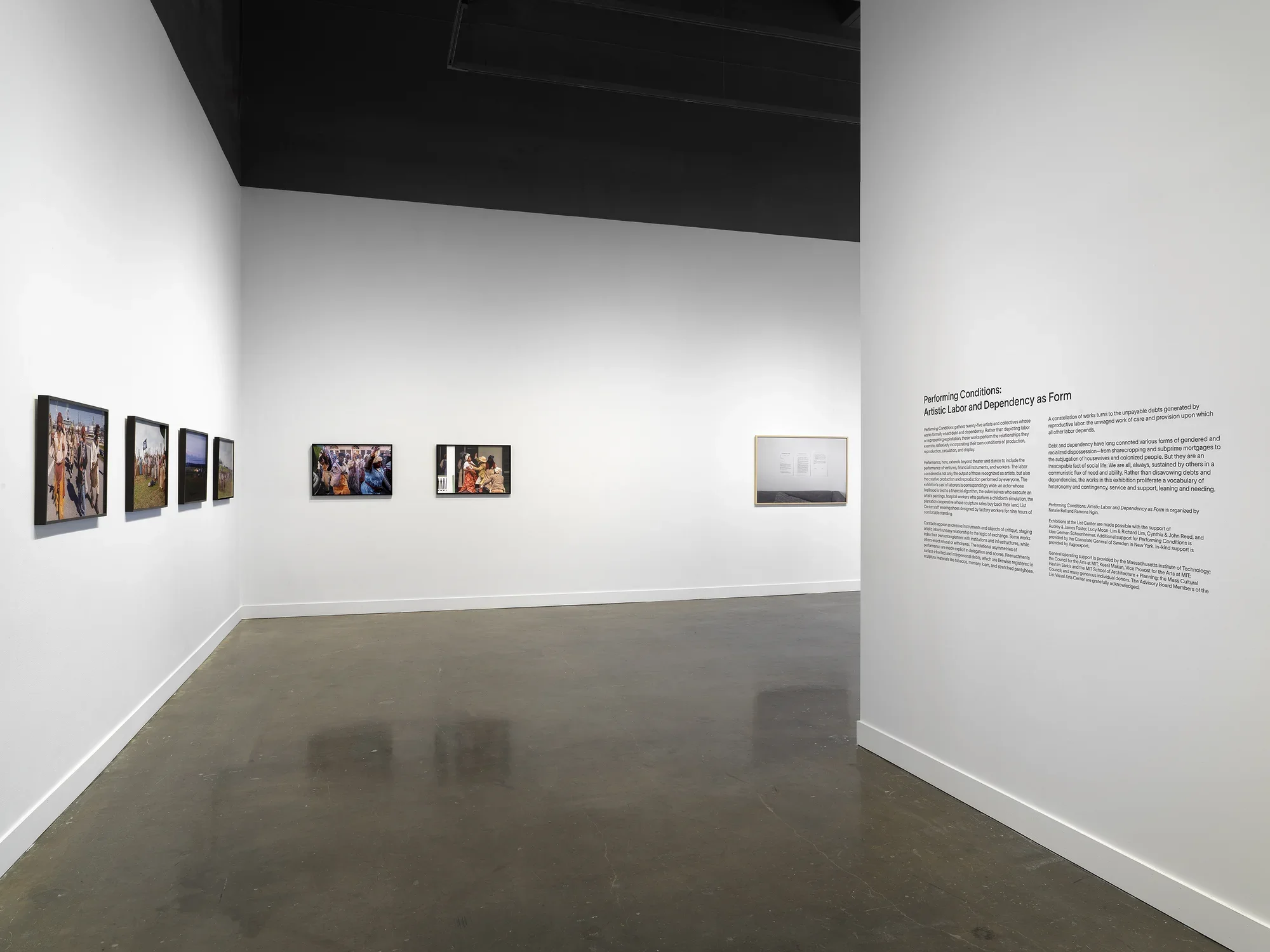 A view of a passageway into a gallery. On the left, six framed photographs showing figures in period dress hang on the wall; on the right is a vinyl introductory exhibition text. 