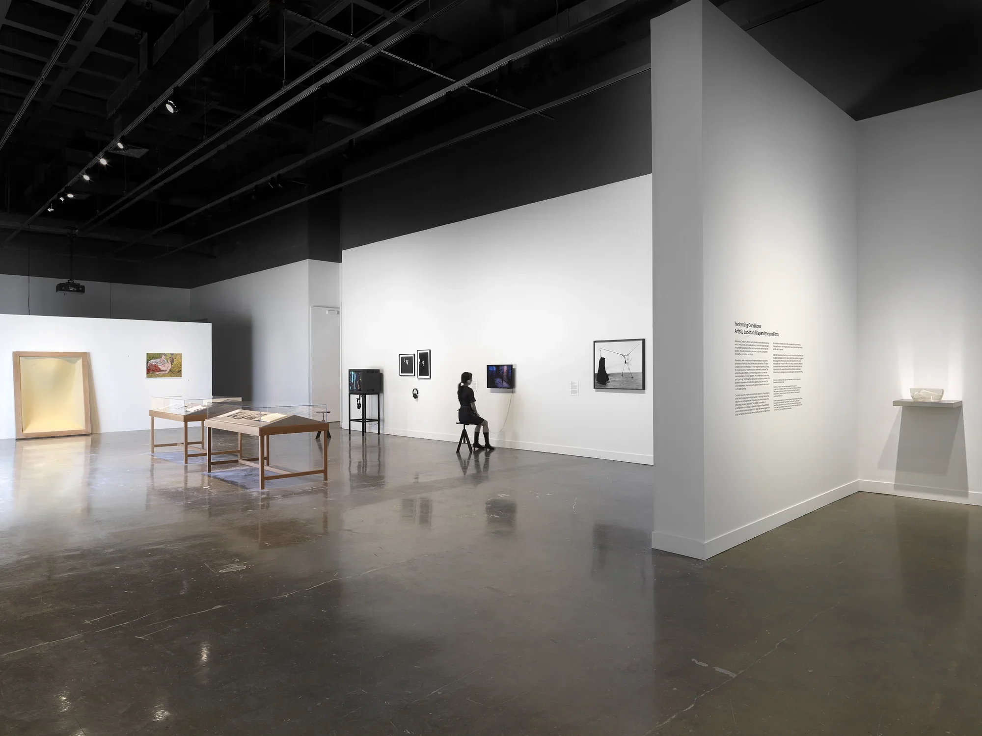 Artworks fill a gallery space, including photographs in freestanding wooden vitrines, a small glass sculpture on a metal wall shelf, video monitors with headphones, framed grayscale photographs of performances, and a yellow rectangular foam sculpture leaning against a wall. 
