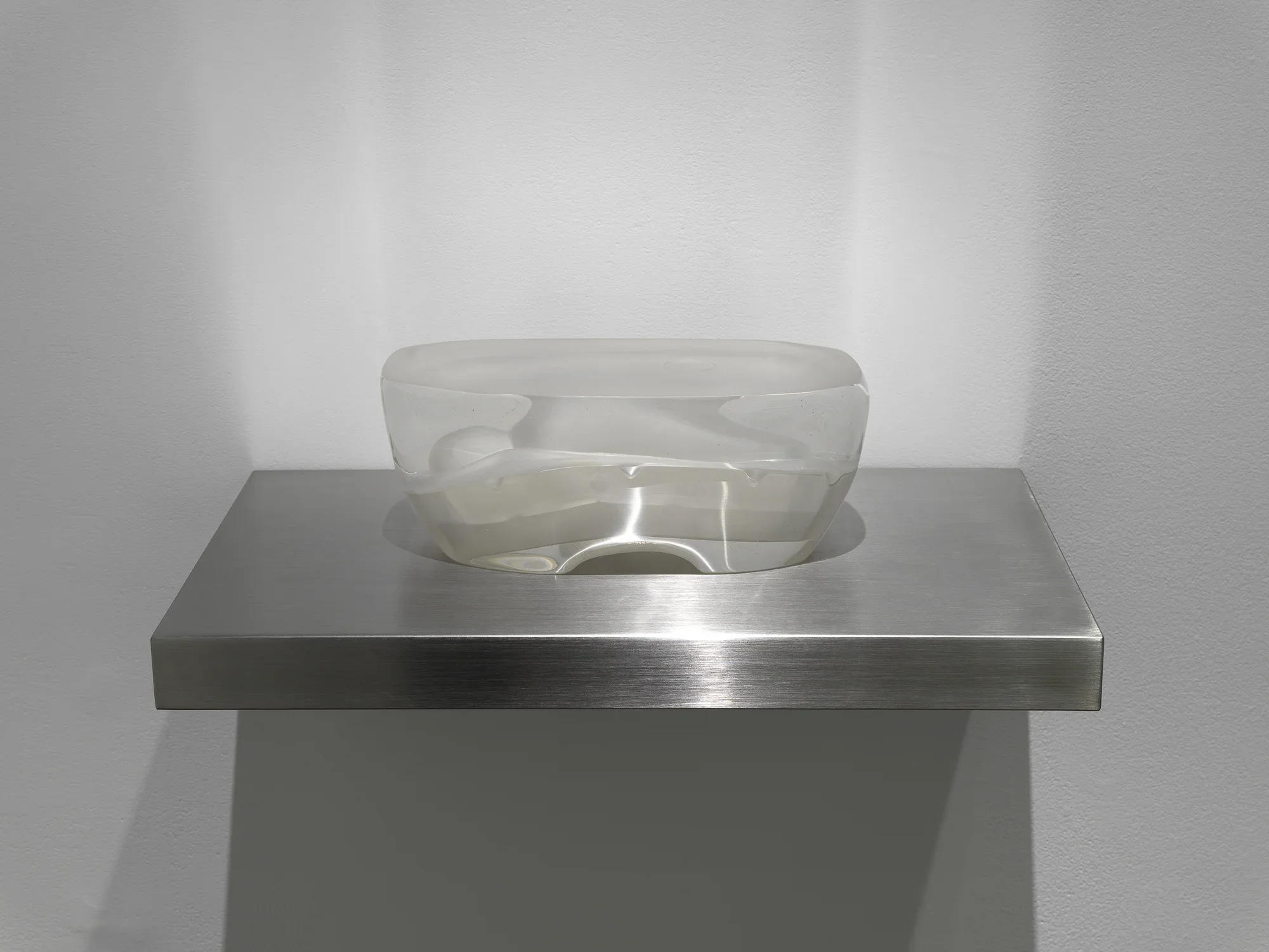 A vaguely boat- or wonton-shaped transparent glass sculpture sits on a metal wall shelf. The sculpture contains an inner space or shape that resembles an abstracted figure, with a rounded head and ovular body.