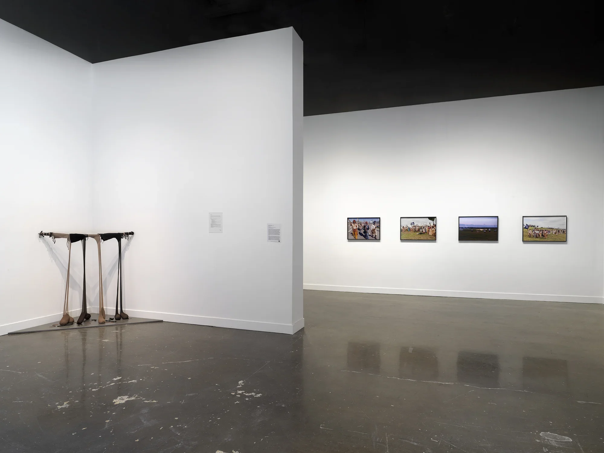 In a corner on the left, a droopy sculpture consists of paired ovular black and brown masses hanging from stick wedged horizontally between the walls. On the right, on a wall further back, framed photographs depict a procession of people carrying flags and weapons. In the center, a sheet of paper with text hangs on the wall.