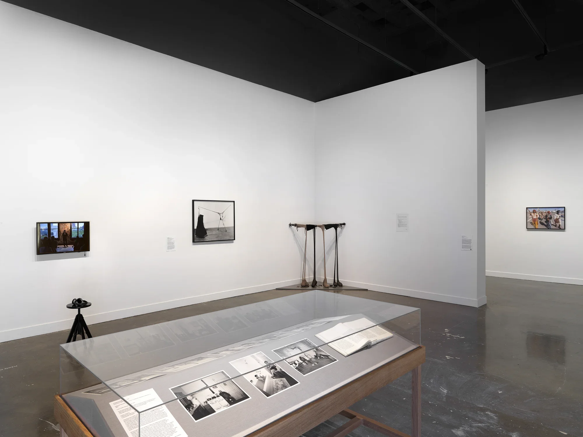 In the foreground, grayscale photographs and an open book occupy a wooden vitrine. The photographs show people in interior spaces, like a man sitting in a kitchen near a stovepipe and a child in a bath near his mother. Other artworks fill the background, including a droopy sculpture, a video monitor depicting a lecture, and a photograph of a procession.