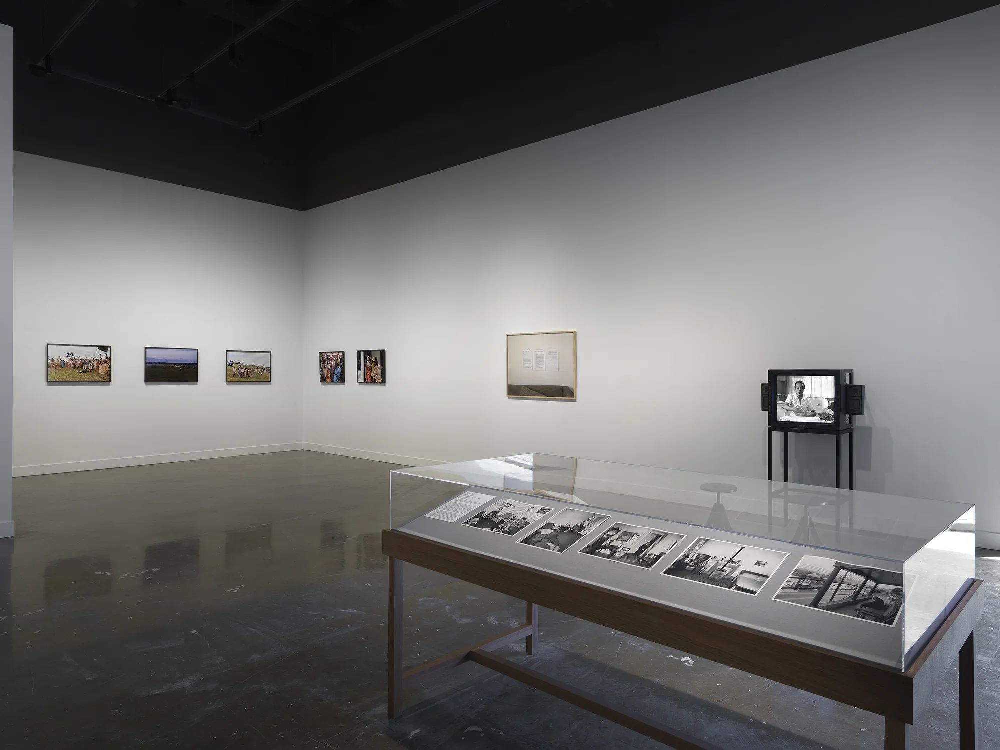 In the foreground on the right, a freestanding wooden vitrine is filled with grayscale photographs: these include a figure sprawled on a chair and a man sitting limply on a porch. Other artworks fill the background, including a boxy monitor depicting a seated woman and a framed photograph of printed pages of text hanging on a wall.