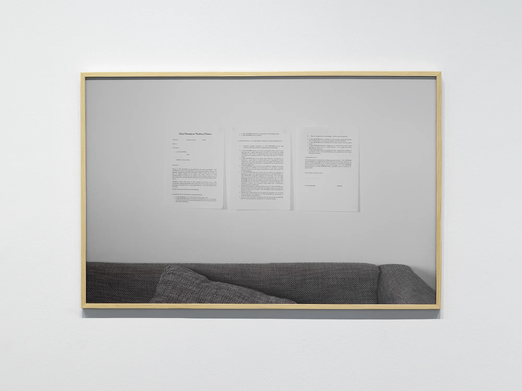 A photograph framed in light wood hangs on the wall. The photograph shows three printed pages hanging, in turn, on a white wall above a couch, only a fragment of which is visible. An unsigned contract is printed on the pages. At the top of the left page, a title is printed in cursive: “Deed of Ownership and Conditions of Existence.”