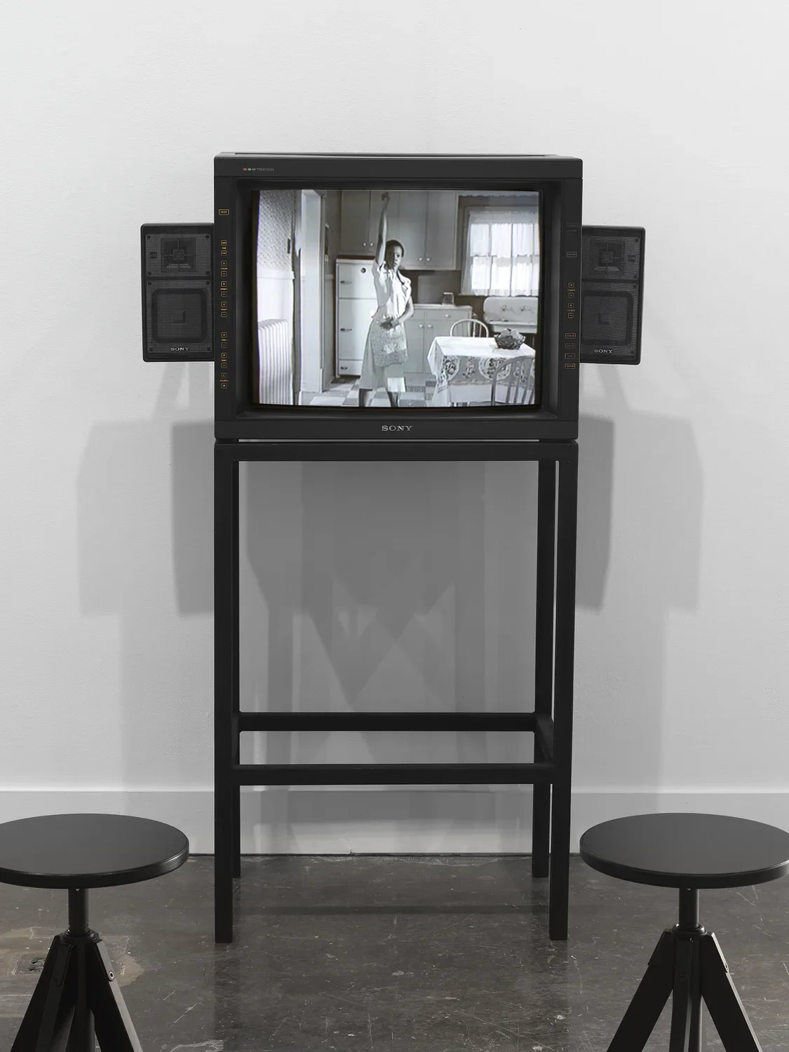 A boxy video monitor with built-in speakers sits on a streamlined metal stand with two stools in front of it. Onscreen, the grayscale image shows a woman wearing white raising her arm straight above her head in a kitchen.