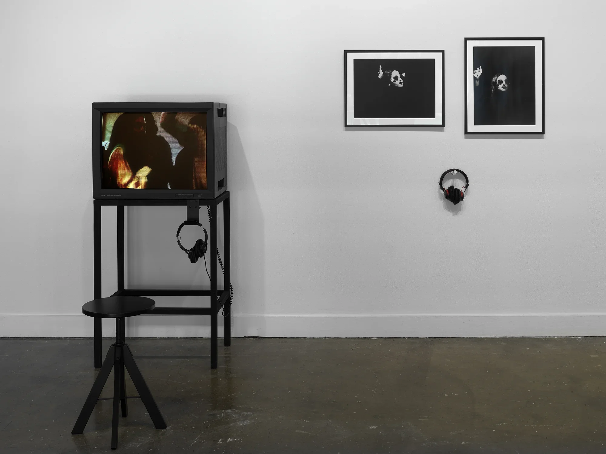 A multimedia installation consists of two parts. On the left, a boxy monitor shows the blurry heads and torsos of dancing figures. On the right, two framed photographs show a woman, her face painted white, wearing sunglasses; little other than her face and right hand are visible. A pair of headphones hangs under the photographs.