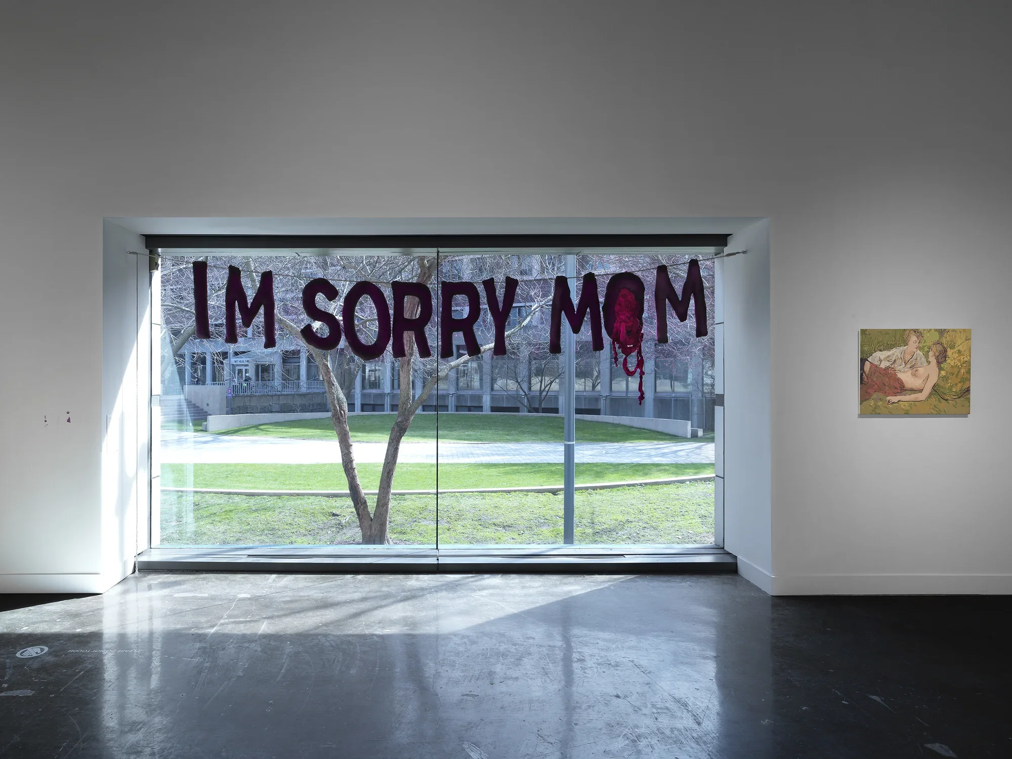 A large window frames a view of a lawn, a building, and a low curving wall. Inside, a sculpture stretches across the upper third of the window: thick, slightly lumpy letters reading “IM SORRY MOM” hang from a taut cable. The center of the second “O” is filled in with tangled red fabric, like viscera. The wall on the left is scuffed; on the right, a small painting showing two women, one topless, hangs.