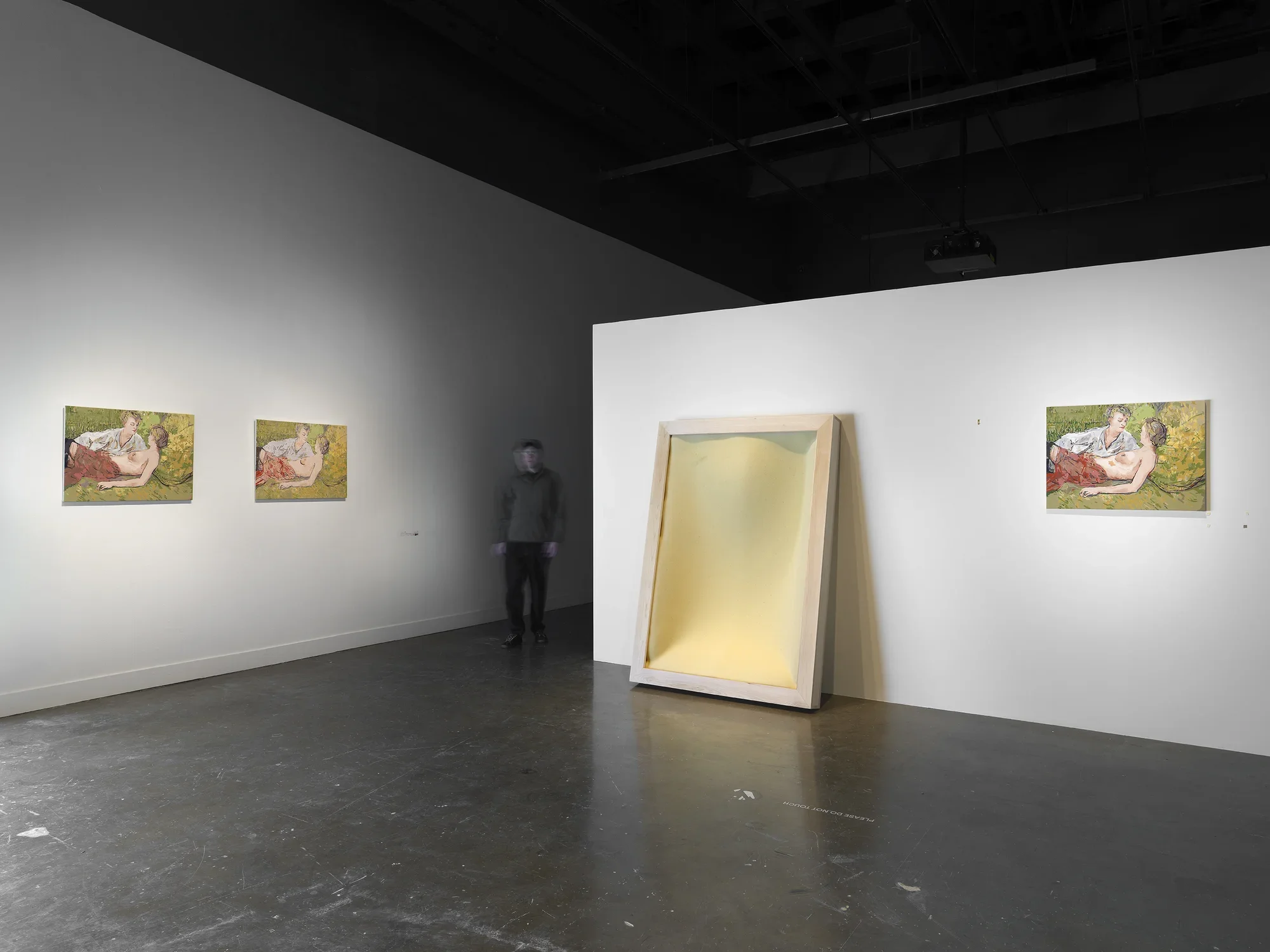 Three nearly identical paintings hang across two gallery walls, which are perpendicular but separated by a passageway traversed by a blurry figure. The golden-green paintings show two women reclining: The one in the foreground is topless, and seems almost cradled by her clothed companion, who leans over her. The trio of paintings is interrupted by an open rectilinear wood frame encasing soft yellow foam and leaning against the right wall, which is freestanding and doesn’t meet the ceiling.