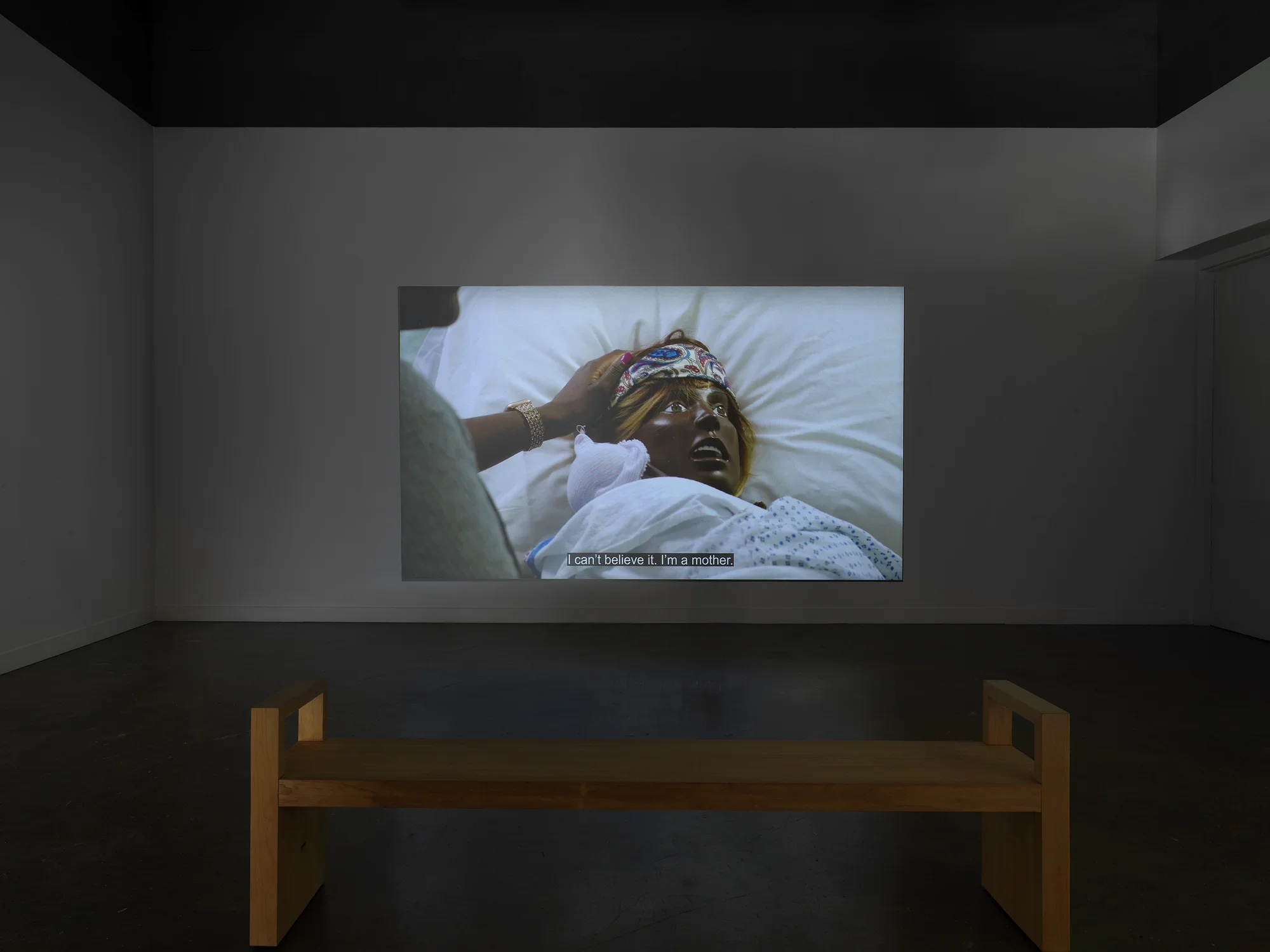 A film is projected in a darkened gallery with a wooden bench. The image shows the head and shoulders of a life-sized mannequin resting on a bed; she has dark skin, blonde hair, and open eyes and mouth. A real person’s hand emerges from behind a blurry shoulder to rest on the mannequin’s head. Captions on the bottom of the screen read, “I can’t believe it. I’m a mother.”