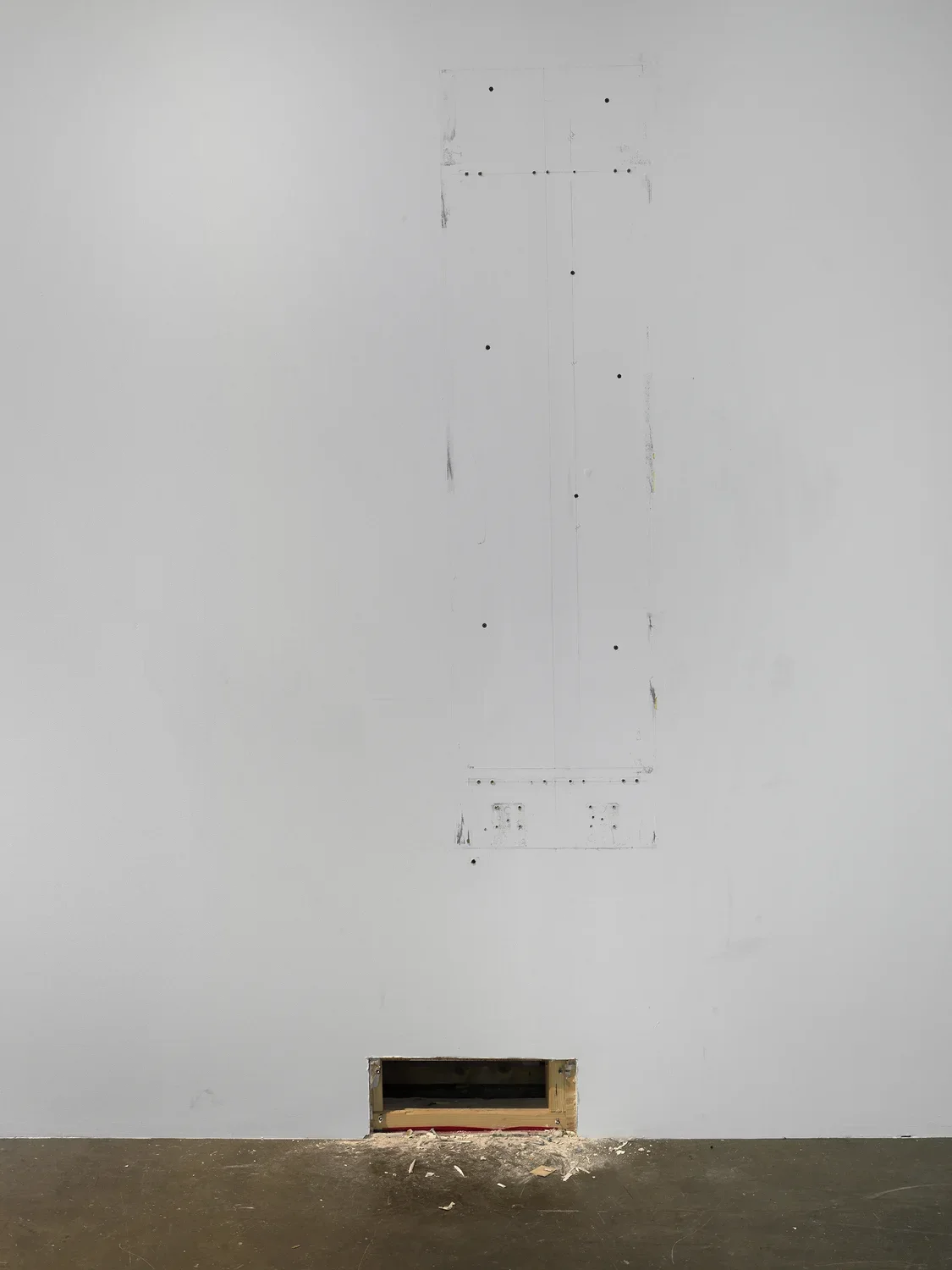 Scuffs and small holes outline an oblong, vertical, rectangular shape on an otherwise bare white wall. Near the floor, a smaller rectangle is cut from the wall entirely, exposing the wooden planks inside. Sawdust and scraps of wood spew from the hole onto the floor nearby.