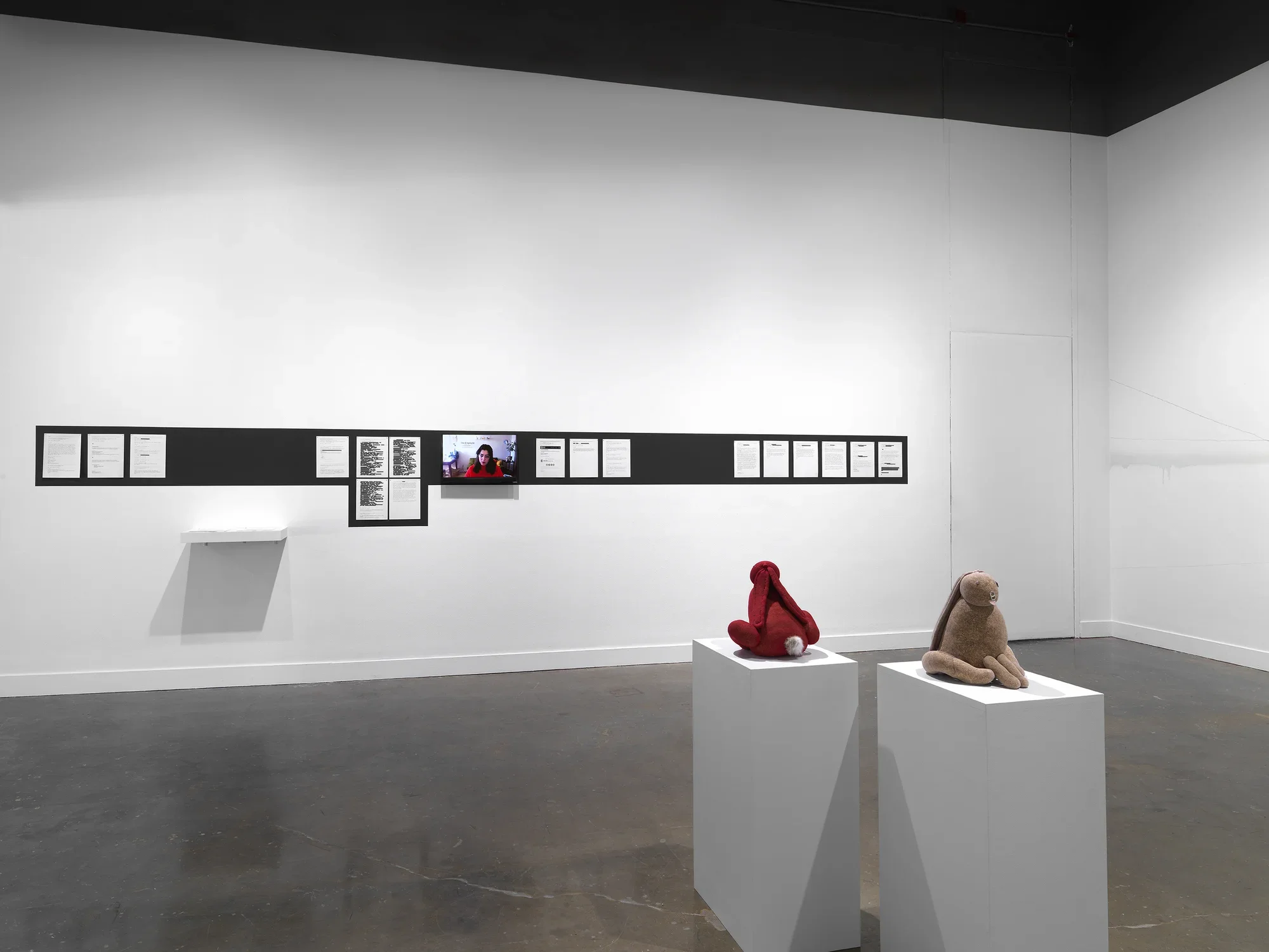 In the foreground, two bunny sculptures—one red, one beige—sit on pedestals, facing away from each other. In the background, on the wall, a stripe of black paint outlines documents tacked in a row (some of their contents redacted in black rectangles) and a video monitor showing a woman’s head. On the far right, a different wall is marked by flowing horizontal lines and a drippy substance.