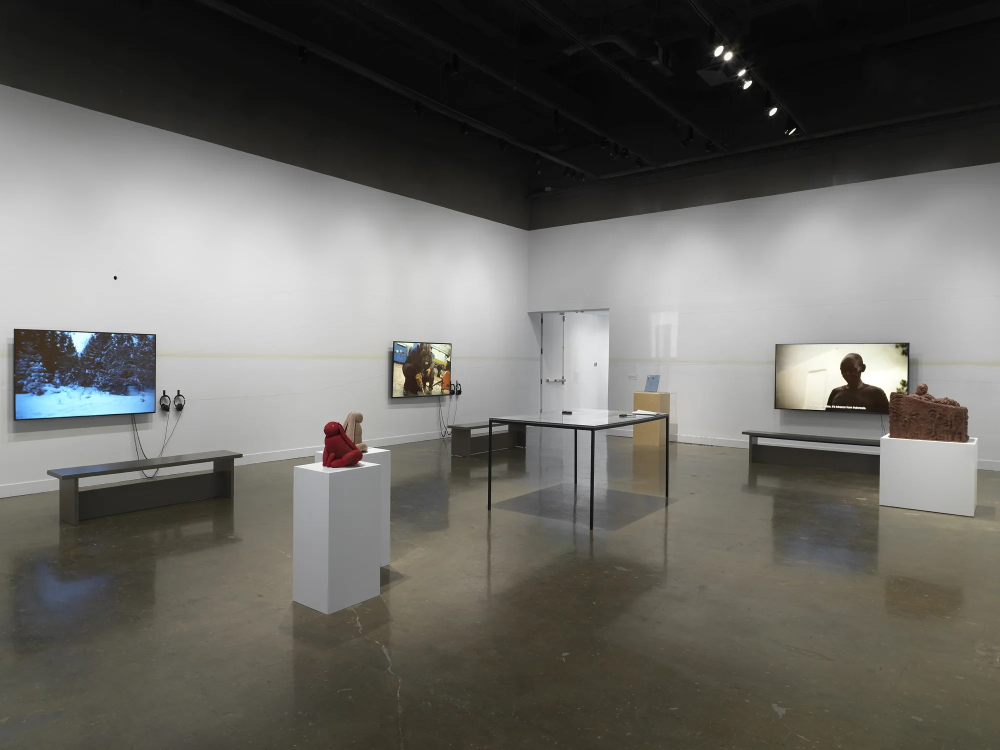 Artworks occupy a gallery, including three large video monitors (each with two pairs of headphones), a table encasing documents, a boxy wooden sculpture, and two soft rabbit sculptures.