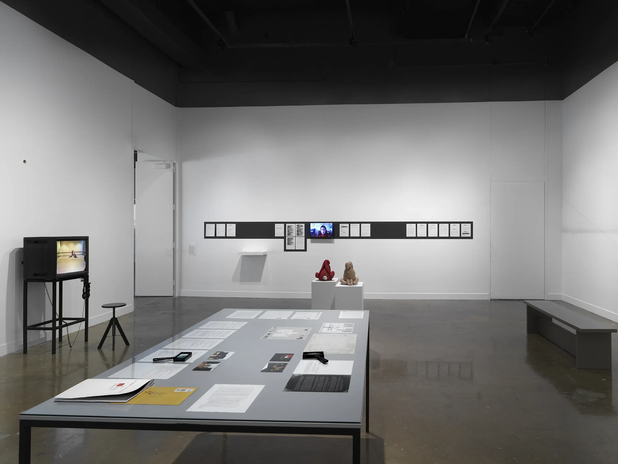 In the foreground, a rectangular table encases documents and drawings—an orange envelope, printed letters, small photographs—under a sheet of clear glass. Handheld magnifiers and a bound volume sit on top of the glass. Other artworks occupy the background, including a boxy monitor showing a dance rehearsal and printed emails hanging in a row on a wall.