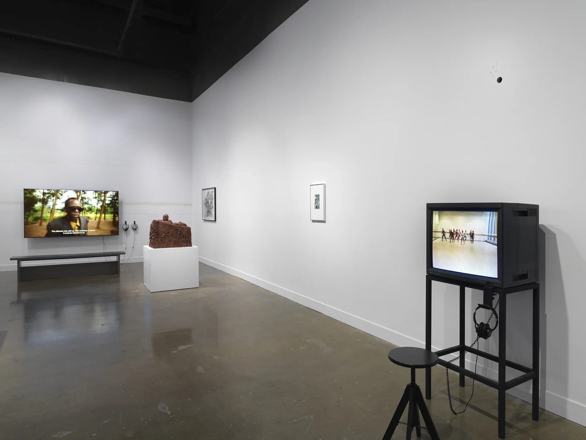 In the foreground on the right, a boxy CRT monitor shows a nondescript rehearsal space with a line of figures toward the back. On the left, other artworks recede into the background, including a small framed print, a wooden sculpture with a head poking above a boxy form, and a large video monitor showing a man in sunglasses.