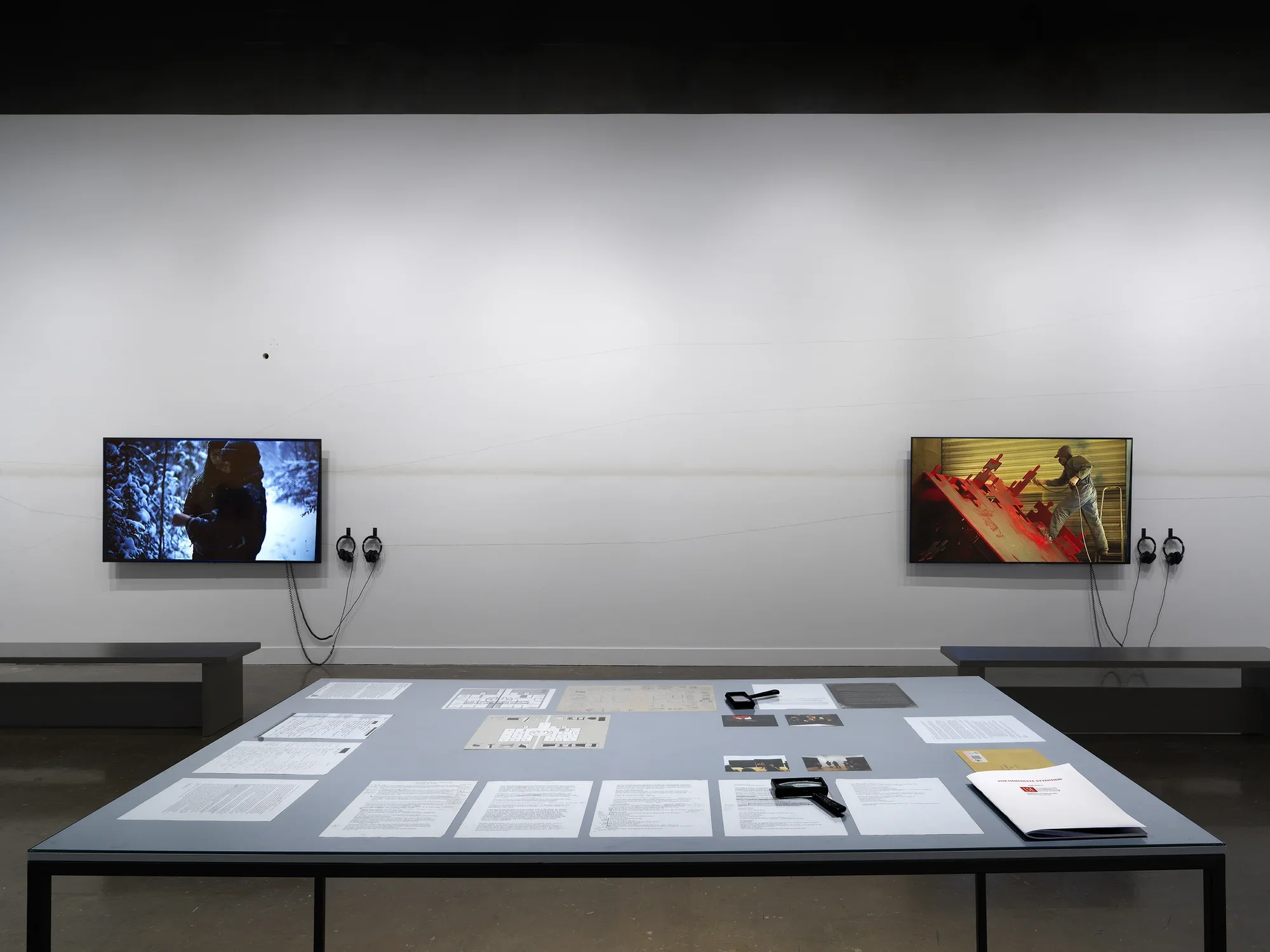 In the foreground, a rectangular table encases documents and drawings—an orange envelope, printed letters, small photographs—under a sheet of clear glass. Handheld magnifiers and a bound volume sit on top of the glass. Two large flatscreen monitors hang on a wall in the background: one shows figures in a snowy landscape; the other shows a factory worker fabricating a red puzzle-like sculpture.