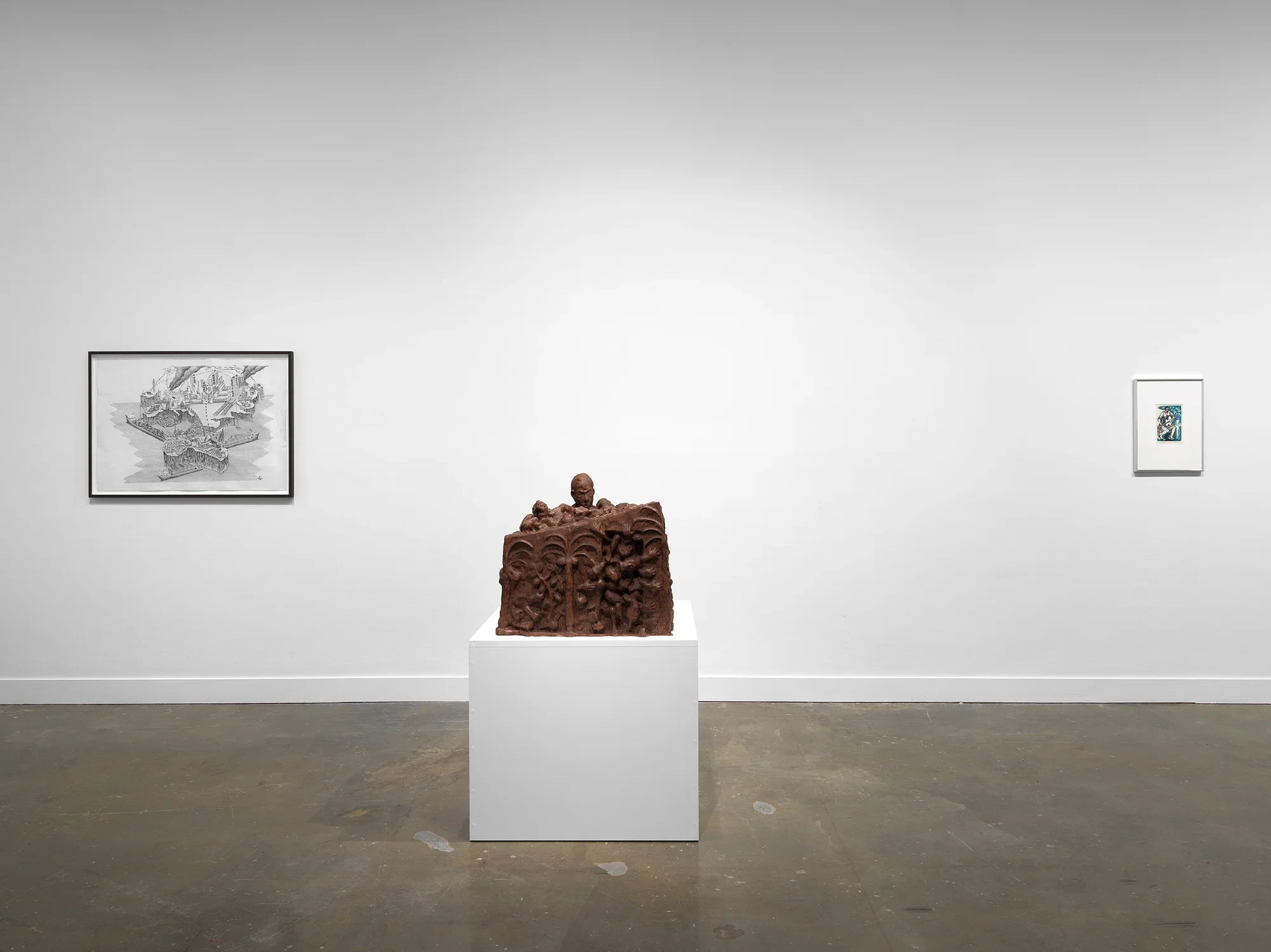 In the center, a boxy wooden sculpture sits on a low pedestal: its front face is carved with palm trees and grasping hands, while a head emerges from the sculpture’s top, looking toward the viewer. Framed works occupy the wall on either side: a black-and-white drawing of an abstract map, and a blue print showing a woman playing guitar.