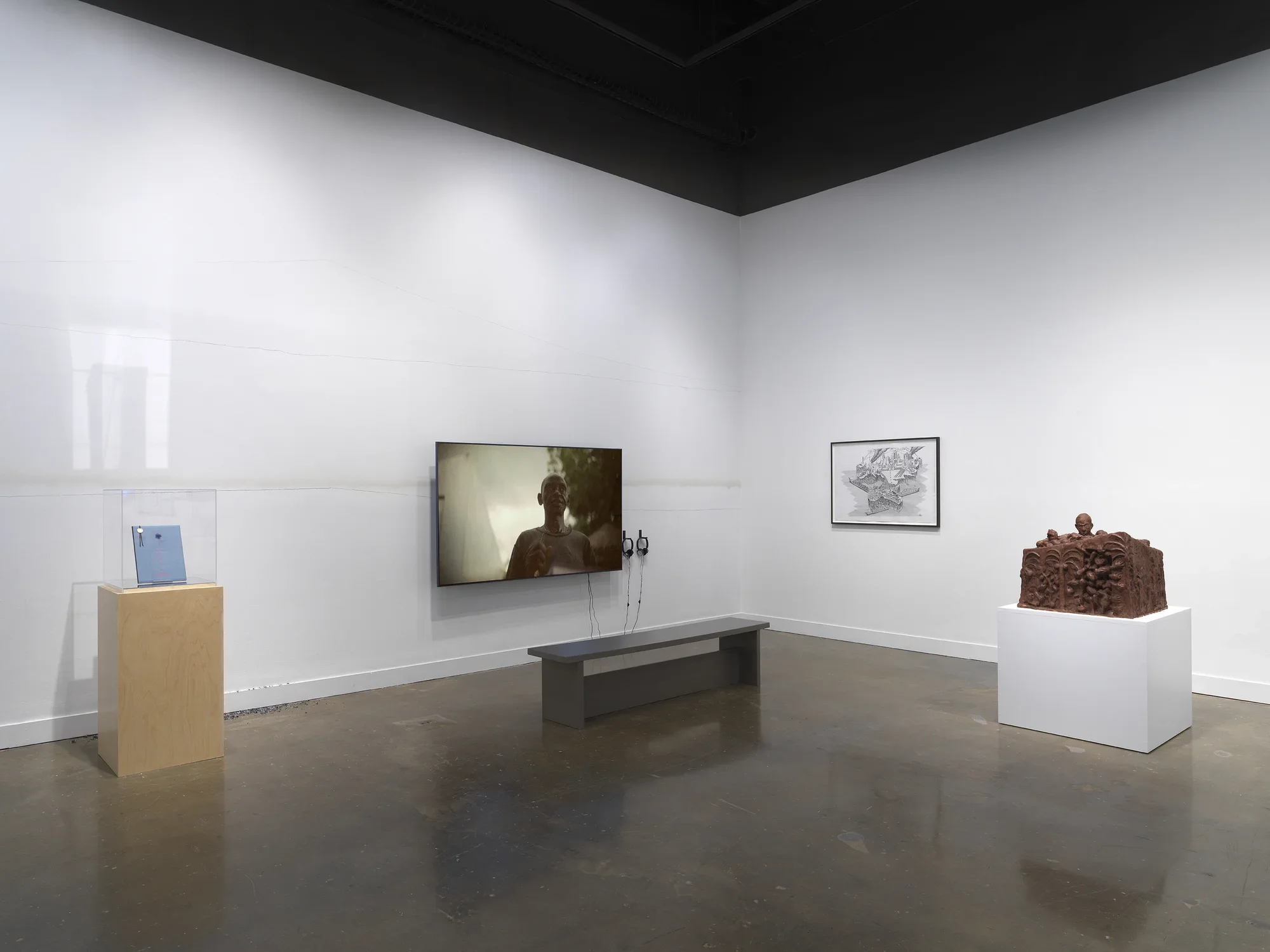 A group of artworks—including a flatscreen monitor, a wooden sculpture, a grayscale drawing, and a blue booklet encased on top of a wooden pedestal—occupy a corner of a gallery. One of the walls is marked by faint, gently sloping horizontal lines.