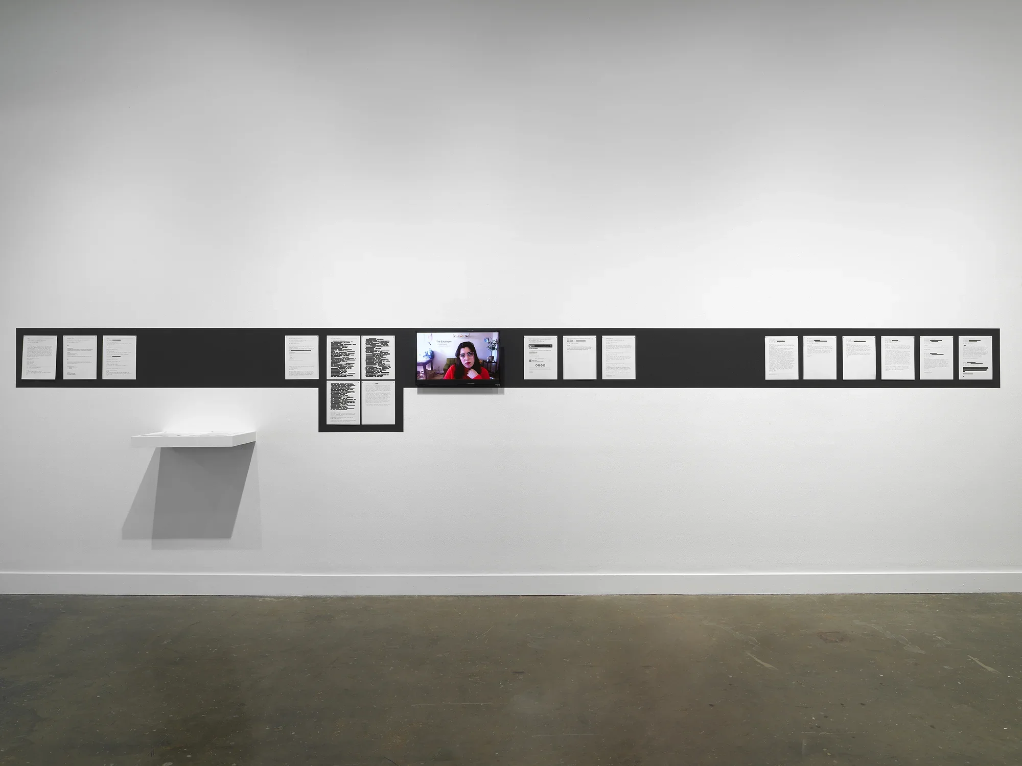 On a white wall, a stripe of black paint outlines printed documents tacked in a row (some of their contents redacted in black rectangles) and a video monitor showing a webcam feed of a woman’s head. A small wall shelf holding two stacks of paper hangs below the stripe, towards the installation’s left side.