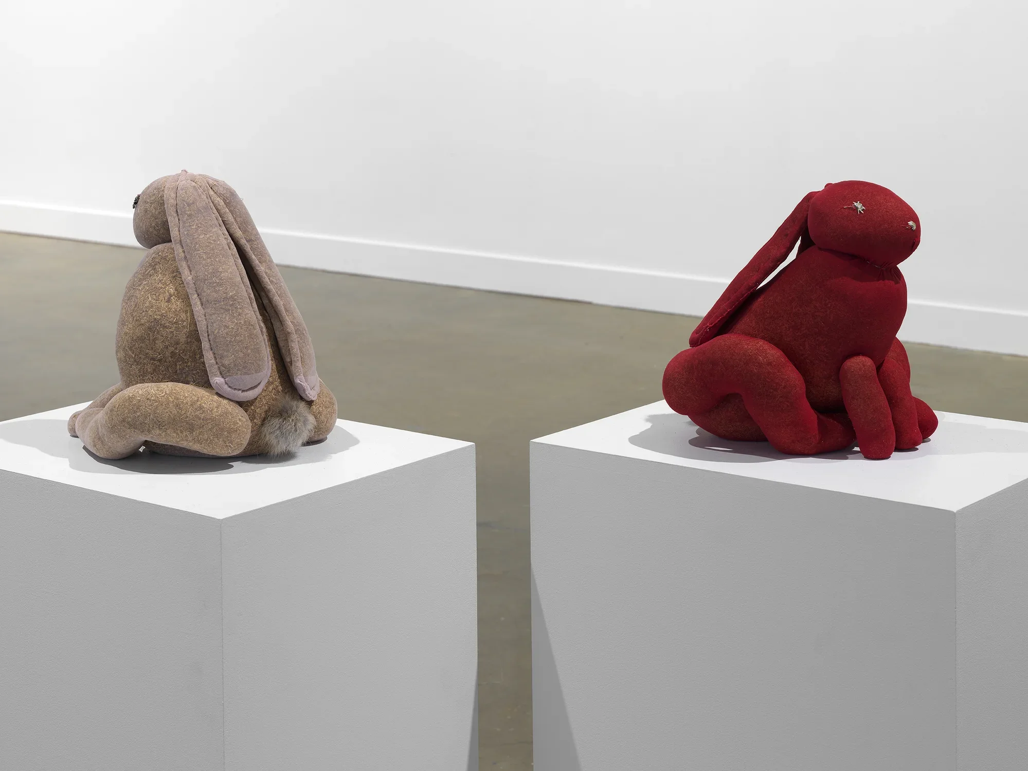 Two lumpy rabbit-shaped sculptures—one brown, one red—are perched on white pedestals, facing away from each other. The soft forms of their bodies—abdomen, head, ears, legs—are made from translucent, delicately sewn pantyhose stuffed with dried tobacco leaves. The red one, on the right, has an eye made from a small white flower.