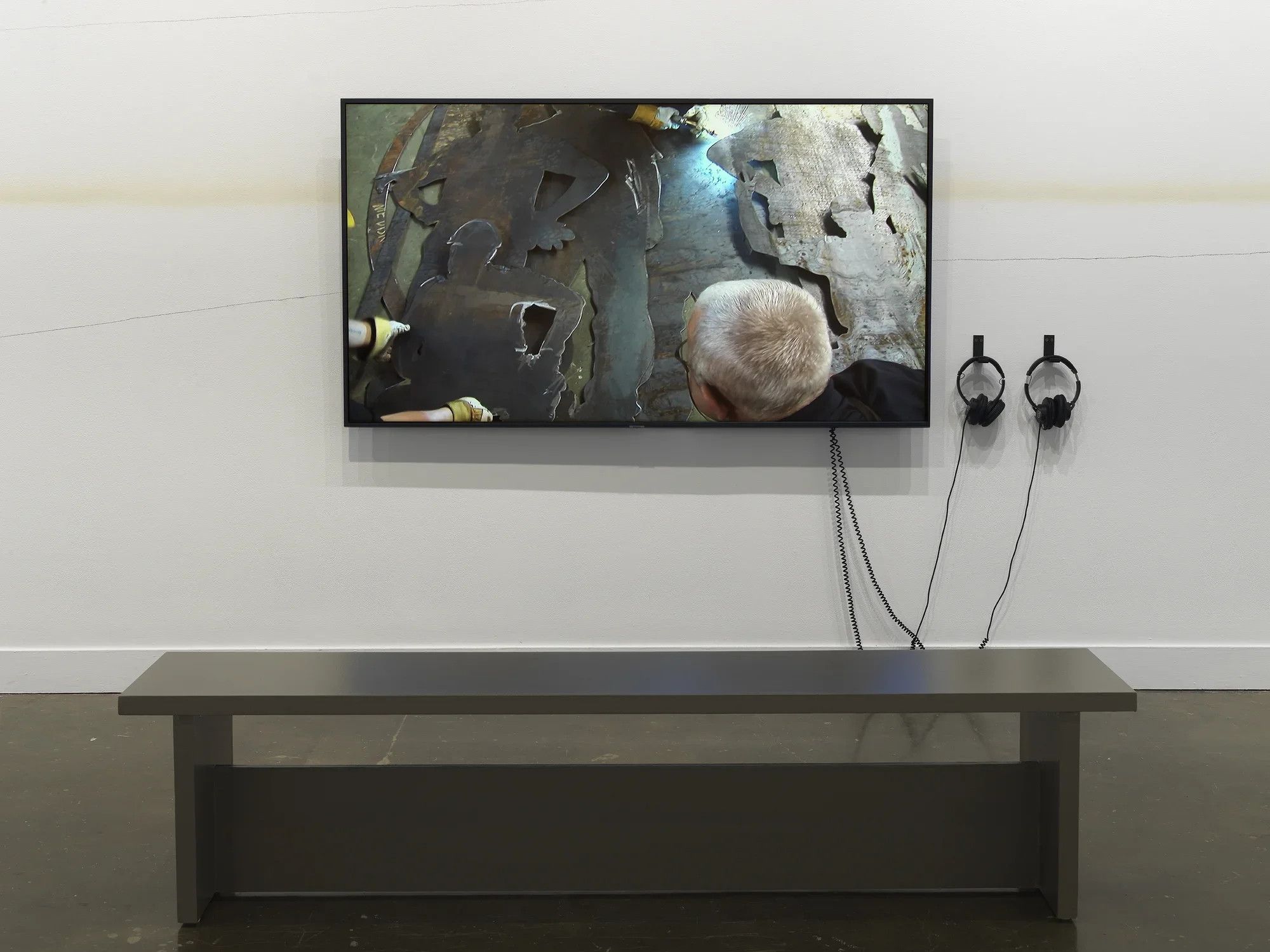A large flatscreen monitor shows the back of a man’s head and layered silhouettes of posing workers cut from silvery metal. Fluctuating lines—two made in pencil, and one diffuse line made of spray tan—travel across the wall behind the monitor.