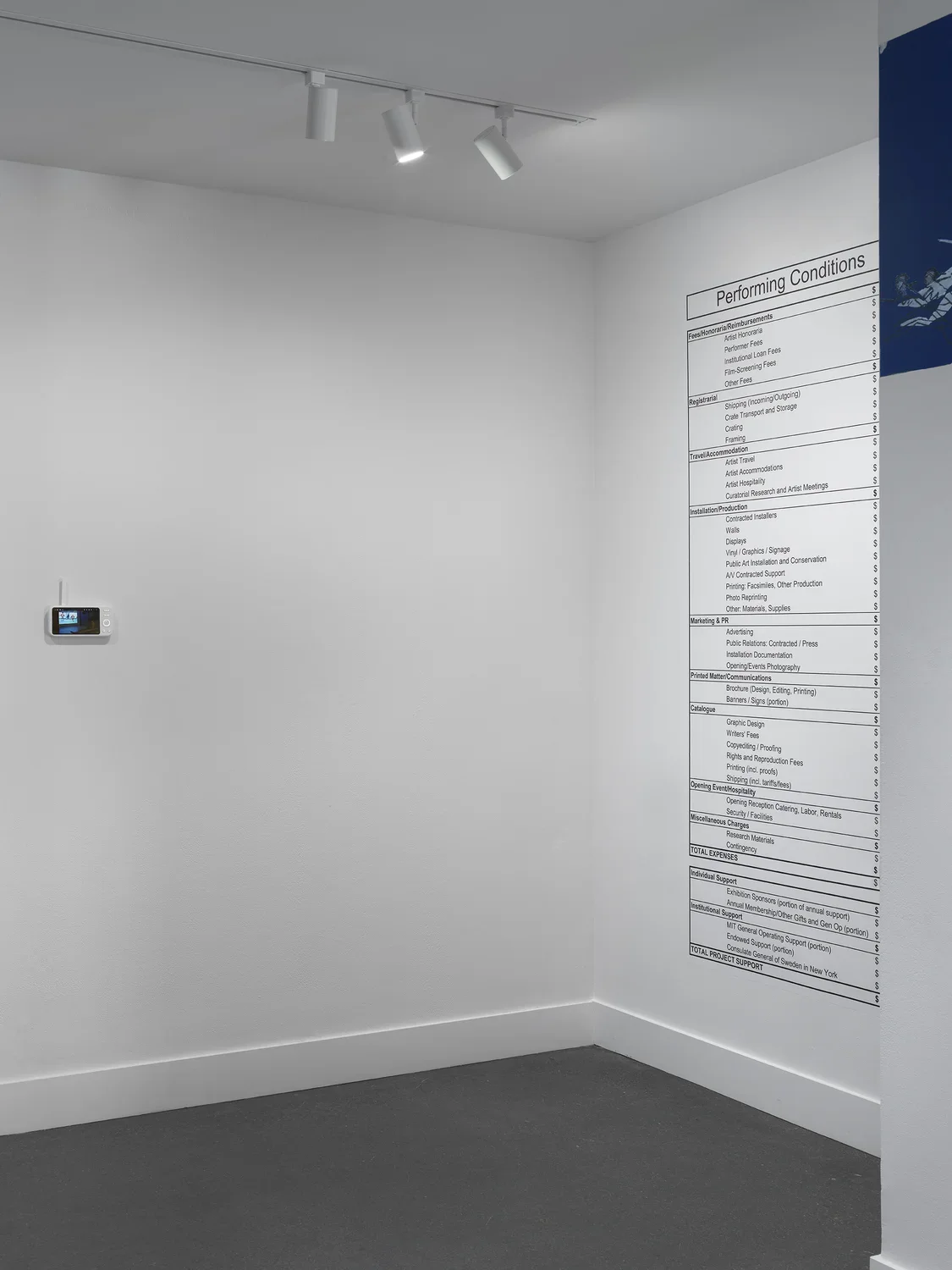 On the left, the screen component of a baby monitor hangs on a wall, dwarfed by the white space around it. On the right, a simple chart labeled “Performing Conditions” is printed on the wall, reaching almost floor to ceiling. The chart is a budget broken up into categories (like Travel/Accomodation and Catalogue) and subcategories (like Writers’ Fees and Shipping).