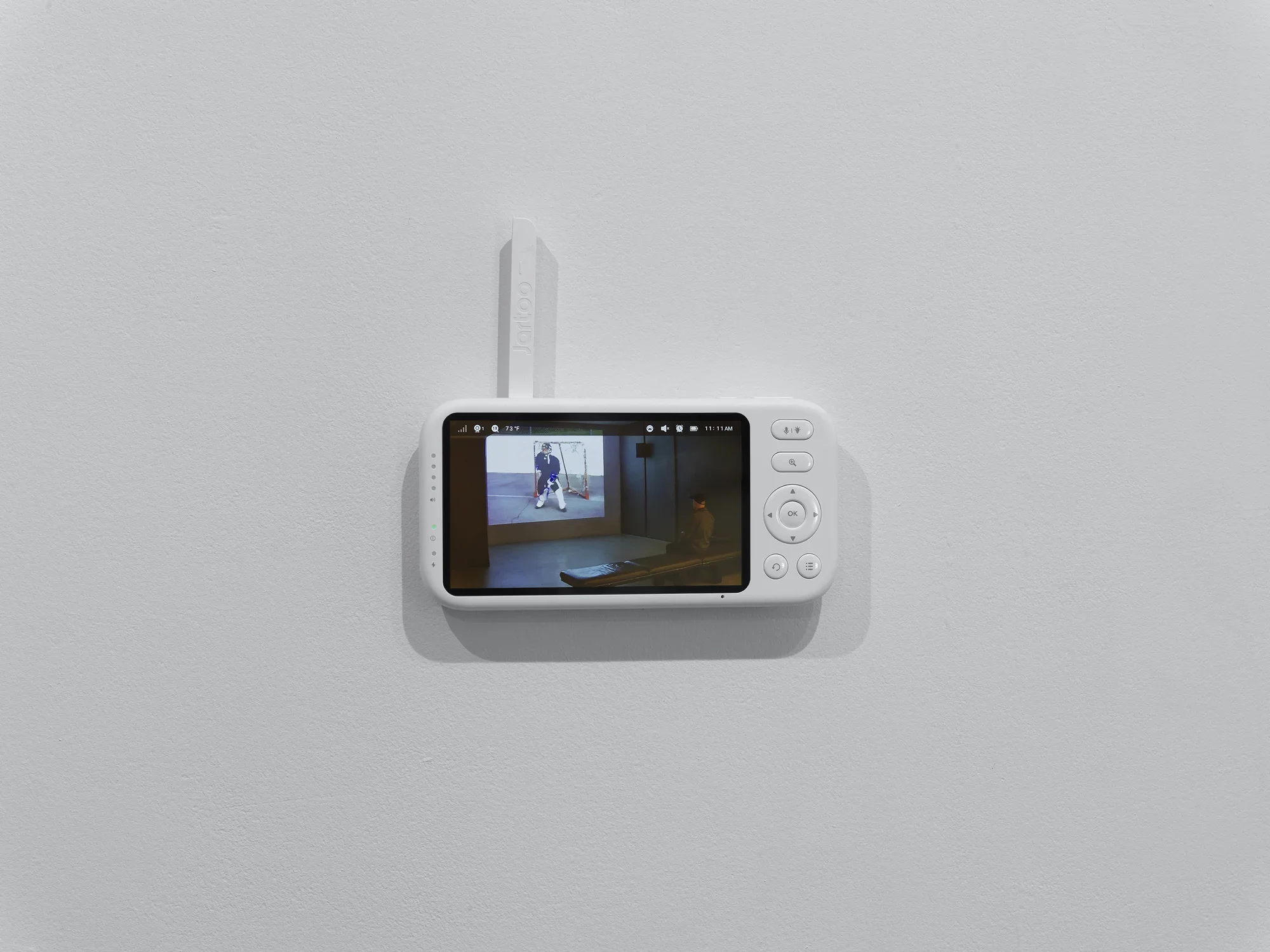 The viewing screen of a baby monitor—with navigation and menu buttons and a white antenna—is attached to a white wall. The feed on the screen shows a person seated on a bench in a dark gallery, watching a projected film.