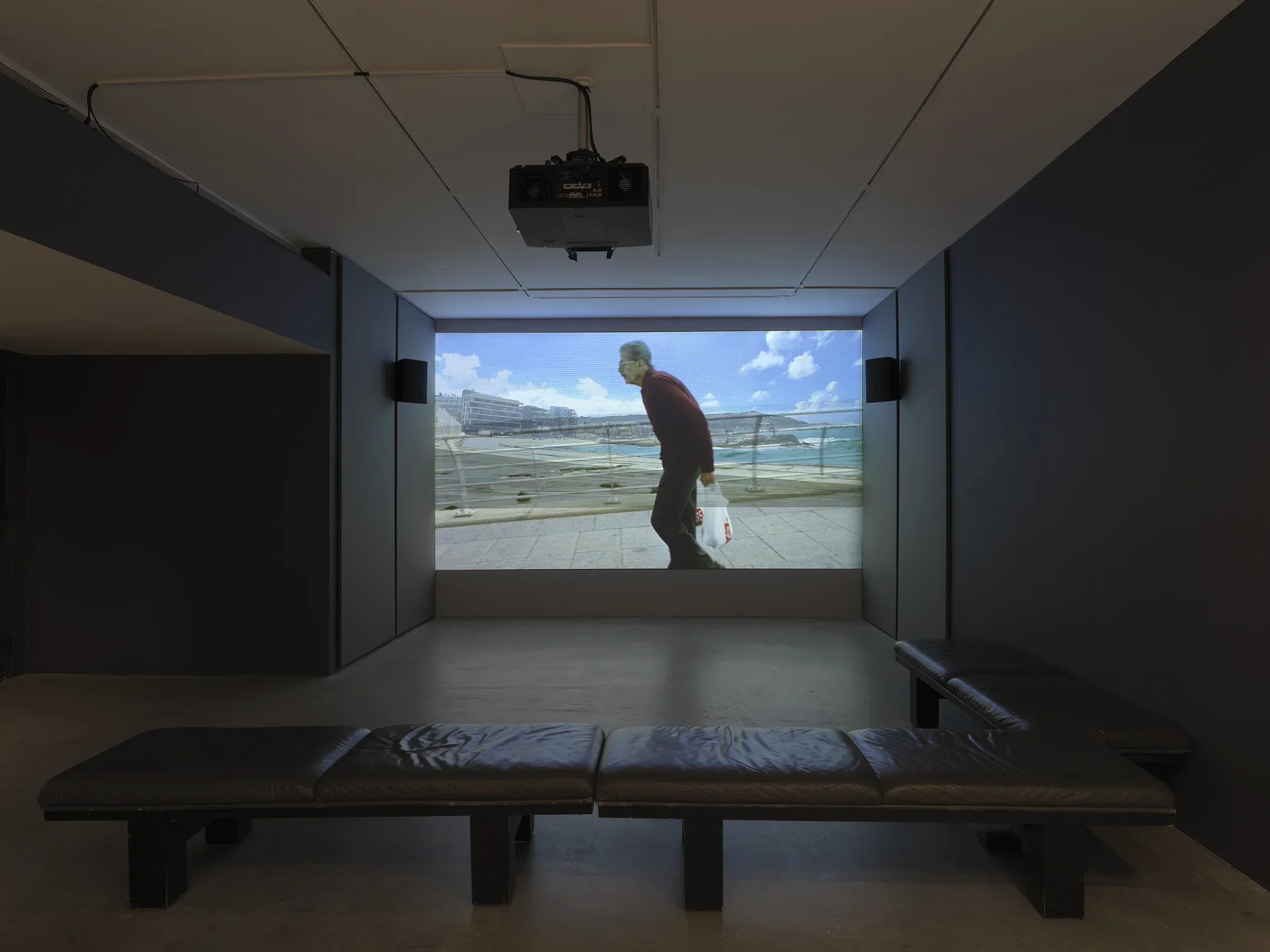 In the background, a film—showing superimposed images of buildings, water, a railing, and an older man—is projected large on a wall, flanked by speakers and acoustic paneling. Three soft benches meet to form an L shape in the foreground.