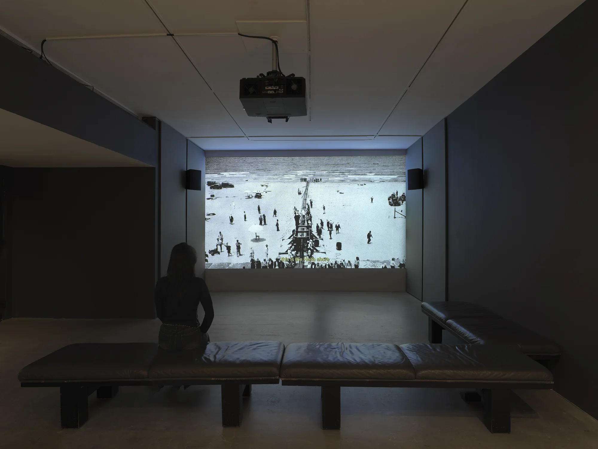 In the background, a film—a grayscale image of a pipeline surrounded by figures, captioned “this is the Haifa shore”—is projected large on a wall, flanked by speakers and acoustic paneling. In the foreground, a person sits on a bench, watching.