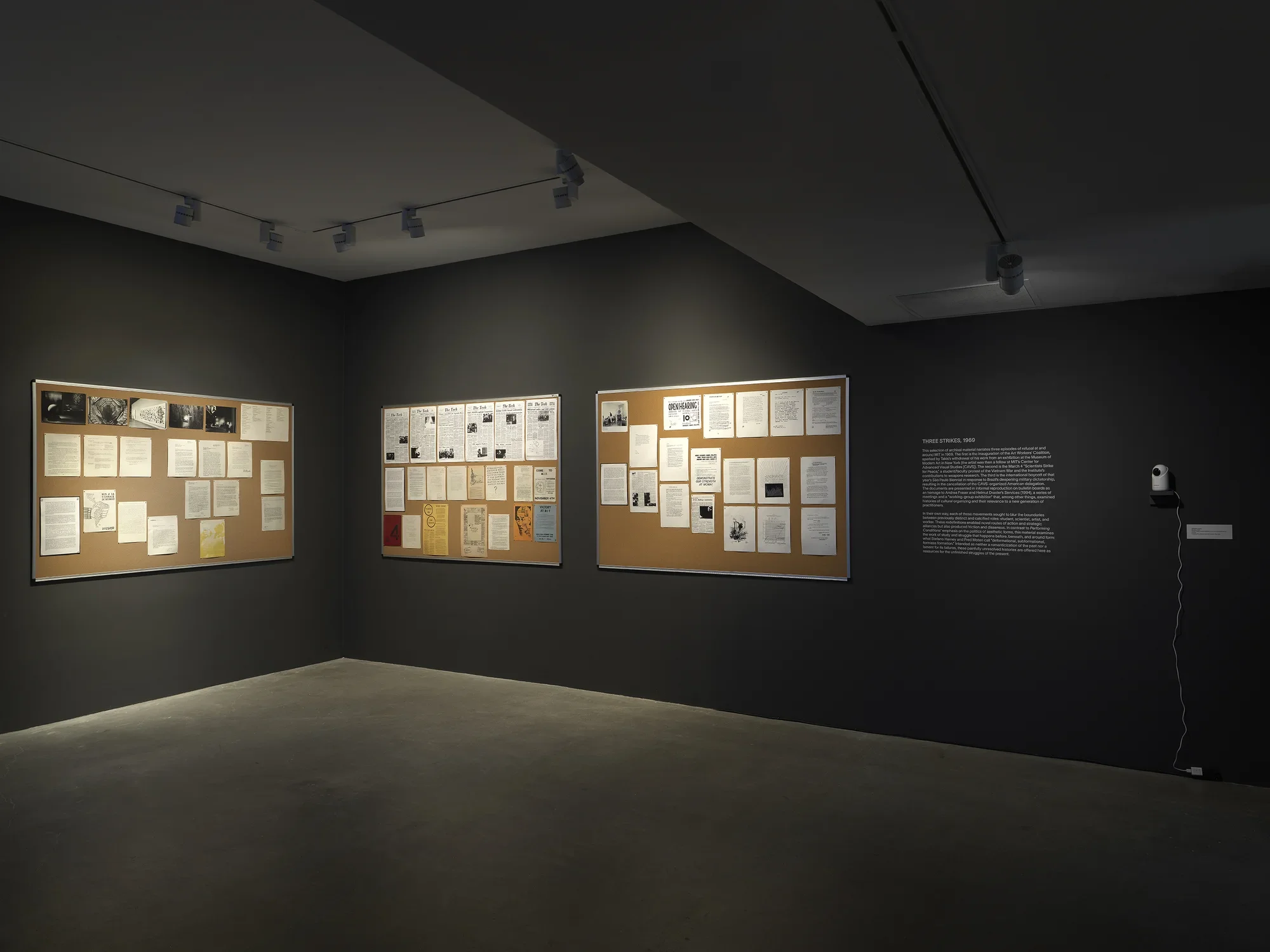 Three bulletin boards covered in reproduced newspaper clippings, photographs, posters, and documents hang in a dark gallery. To their right is vinyl wall text and the camera portion of a baby monitor.