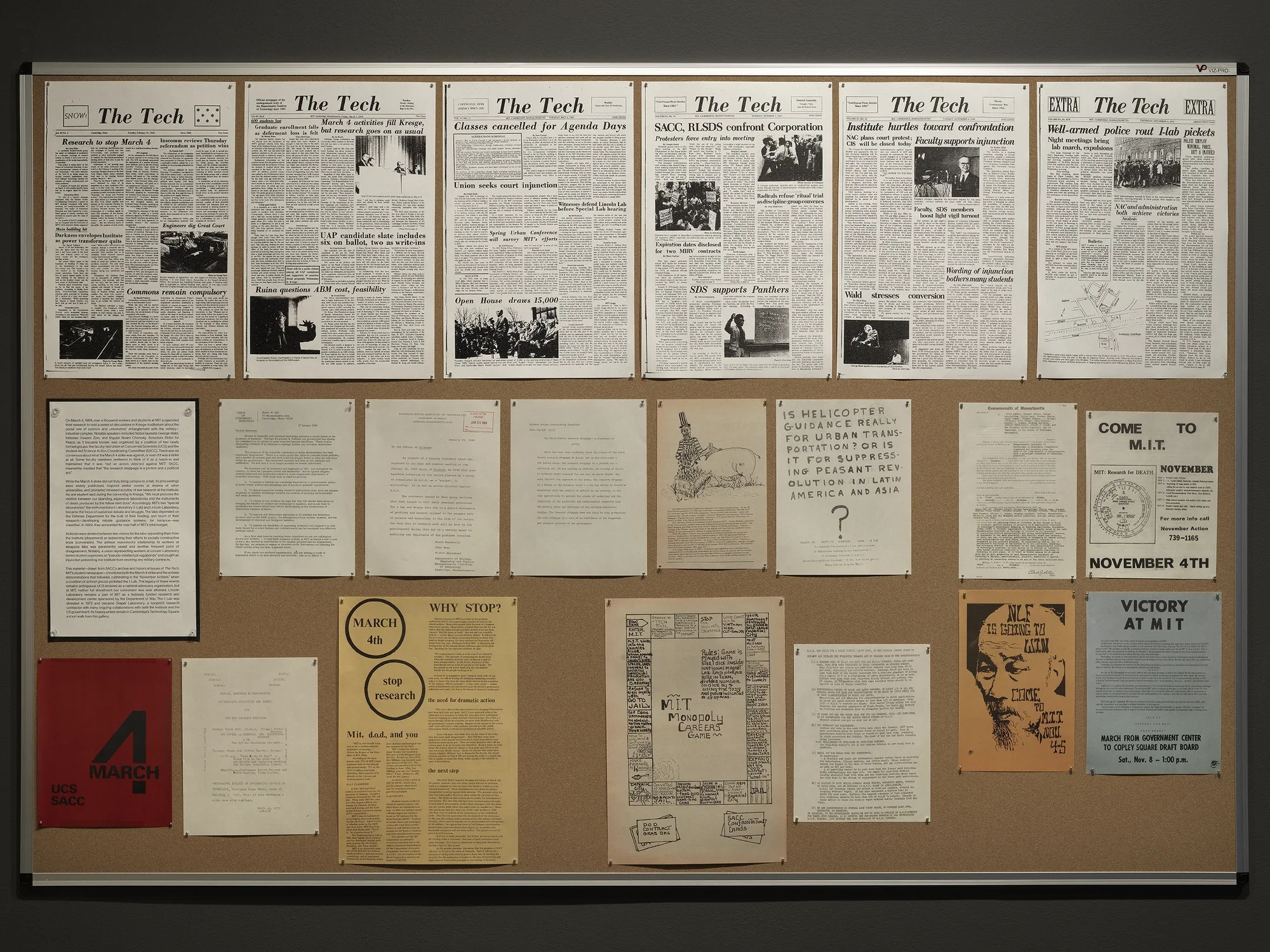 A bulletin board is covered by reproduced documents. In a row at the top, issues of The Tech convey headlines of antiwar protests: “Well-armed police rout I-lab pickets,” reads one. The bulletin board also features copies of posters: one prominently displays the date March 4, while others enjoin readers to come to MIT in November.
