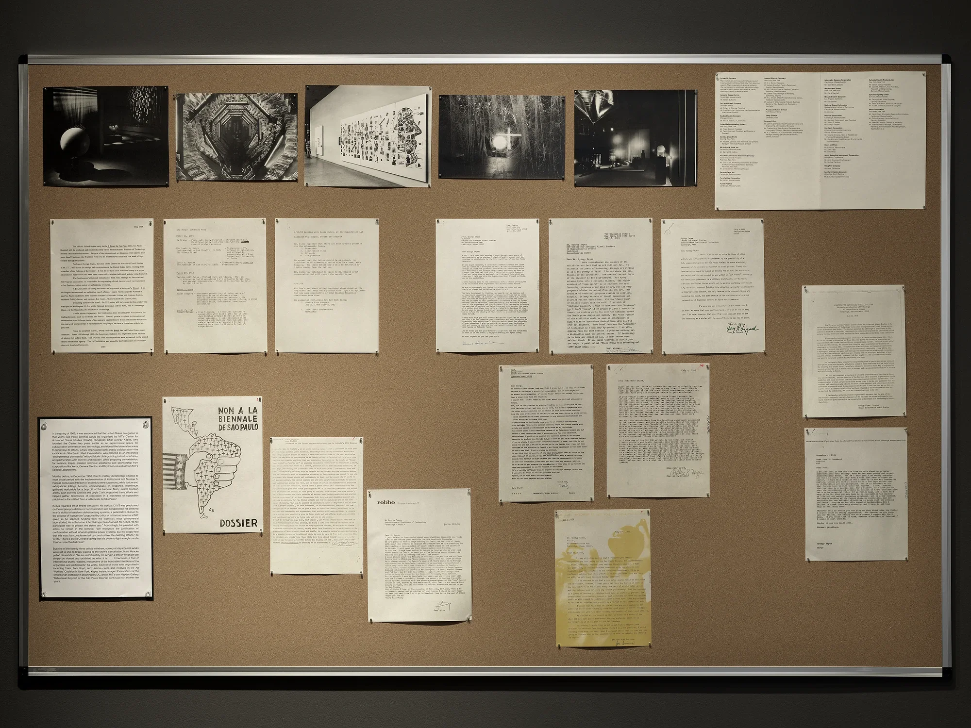 Reproduced documents are arranged on a bulletin board. Most consist of text, but one is a grayscale illustration of the outline of Central and South America, filled in with small crosses. The continent is gripped by a hand with stars and stripes on its cuff. Towards the top of the board, a row of photograph shows an exhibition of technological art: shiny balloons and glowing orbs.