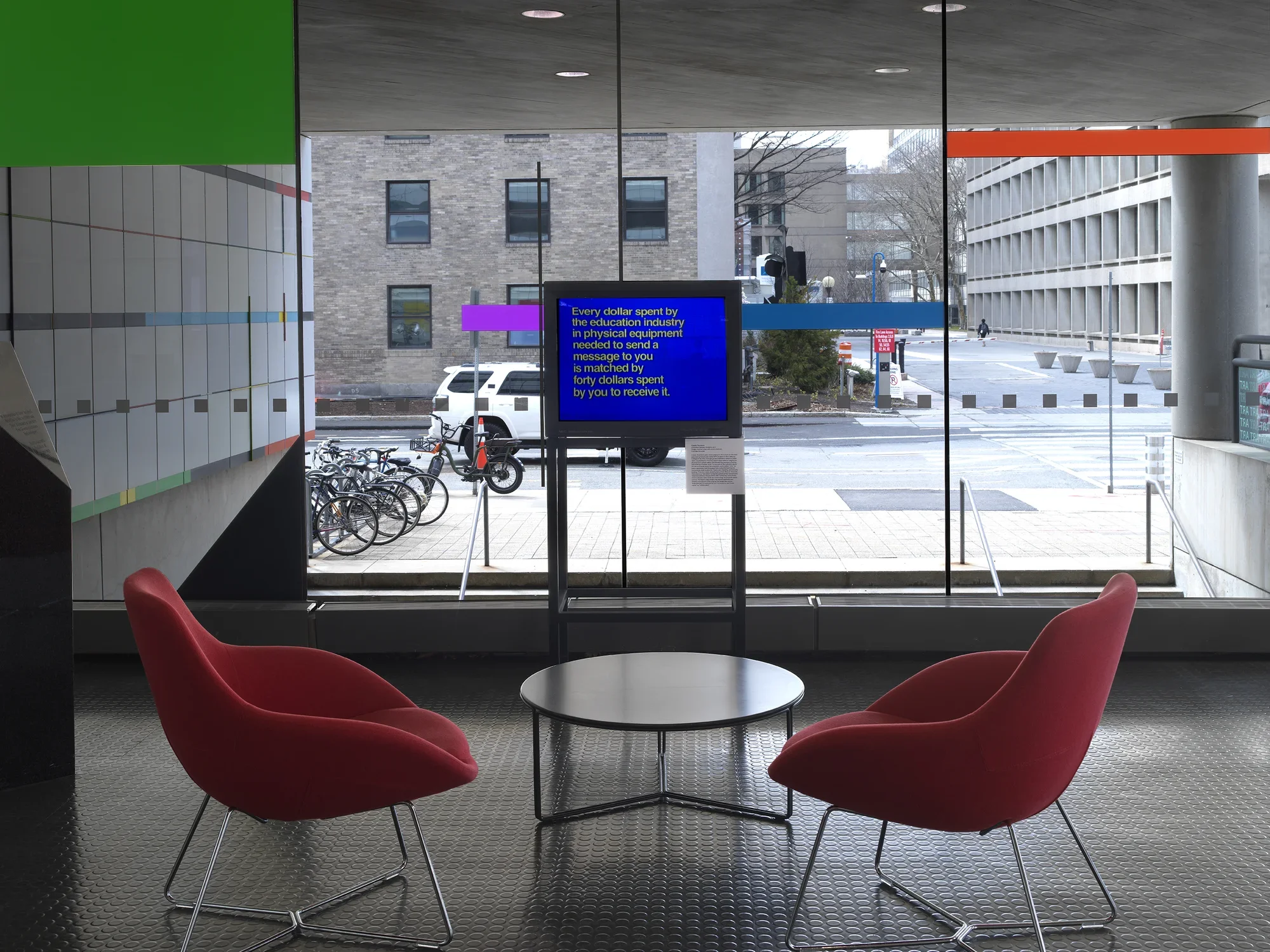 A boxy monitor is positioned against a window, through which a street and the buildings of a campus are visible. The screen shows white text against a bright blue background, reading, “Every dollar spent by the education industry in physical equipment needed to send a message to you is matched by forty dollars spent by you to receive it.” 