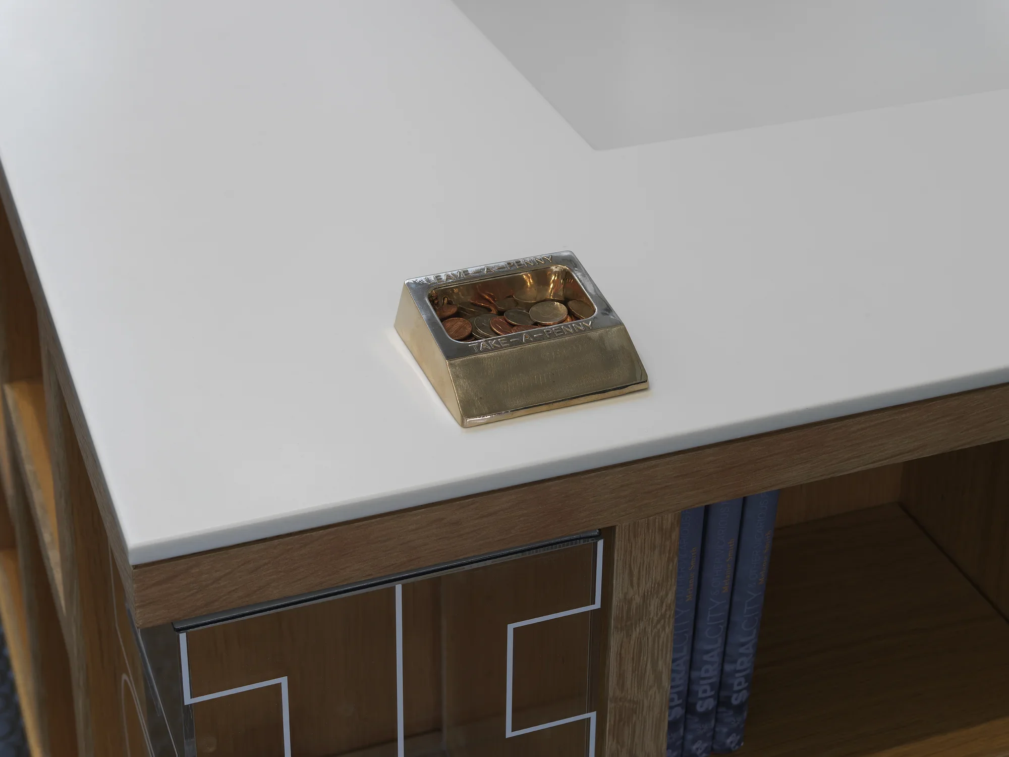 A bronze penny-share dish filled with coins sits on a white desk surface, with concave shelving visible below. The surface of the dish has raised text that reads: “LEAVE-A-PENNY” on its top edge and “TAKE-A-PENNY” on its bottom edge.