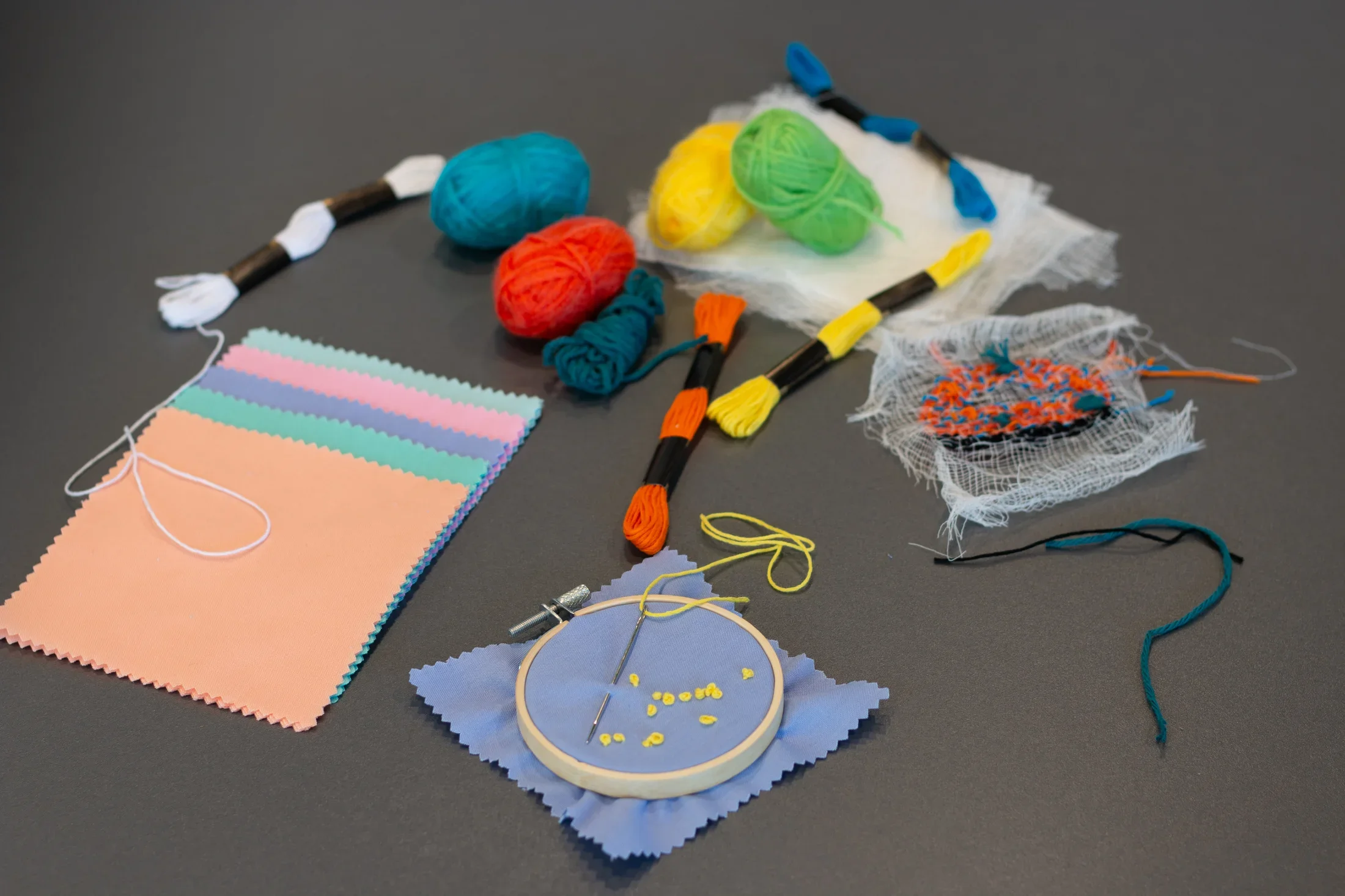 Swatches of fabric, spools of yarn, thread, and periwinkle fabric with yellow french knots in an embroidery hoop are scattered on a gray table.