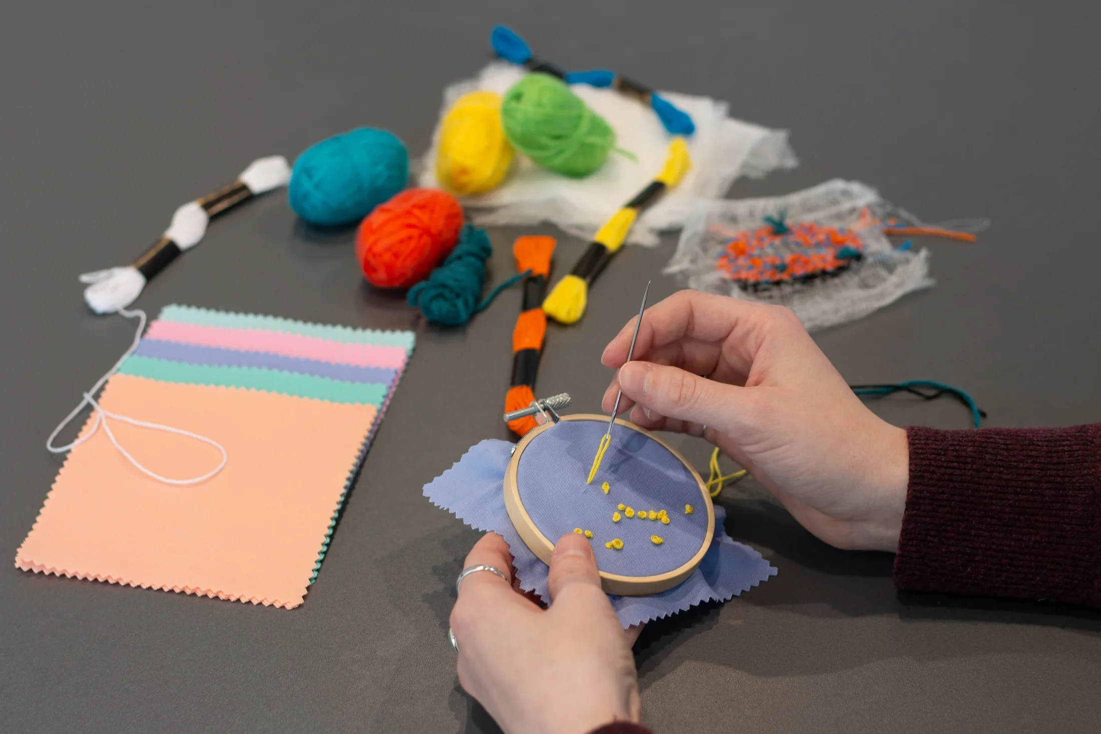 Swatches of fabric, spools of yarn, and thread are scattered on a gray table. Hands sew yellow french knots on a periwinkle fabric swatch in an embroidery hoop.