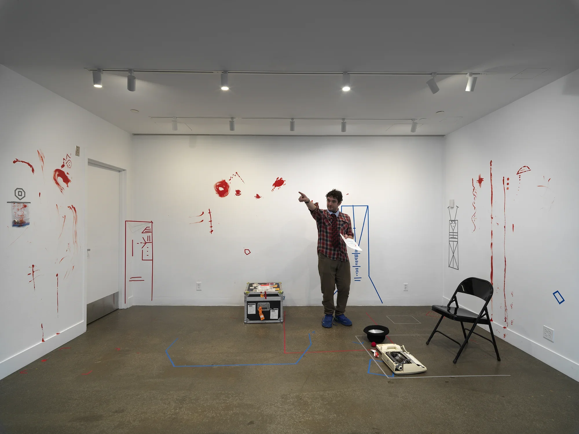 In a gallery, a man wearing a patterned tie holds a printed script in his left hand and points with his right hand. He is surrounded by props and traces of a performance. These include a folding chair, a typewriter, a top hat, and a clown nose on the floor, and obscure geometric figures made of colored tape and more expressive smears of red paint on the wall.