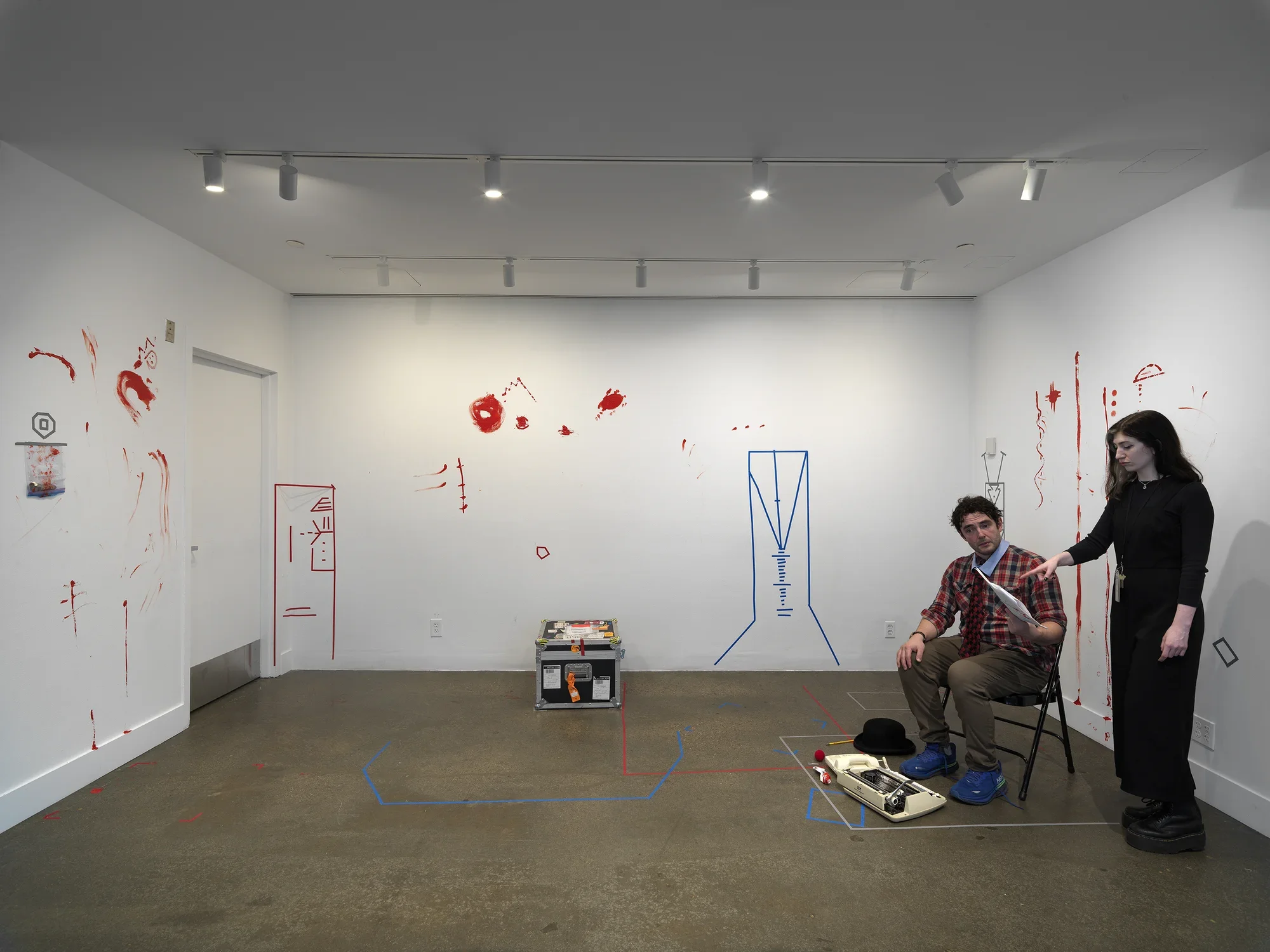 In a gallery, a man wearing a patterned tie sits on a folding metal chair and holds a printed script. A woman dressed in black stands behind him and points to the script. The walls are marked by obscure geometric figures made of colored tape and more expressive smears of red paint.