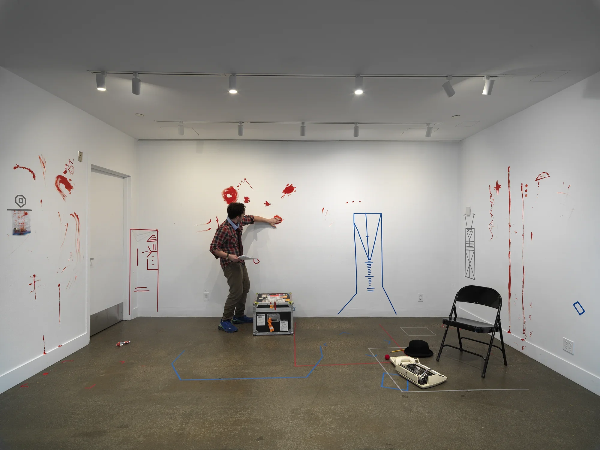 In a gallery, a man wearing a patterned tie holds a printed script in one hand. With the other palm, he smears red paint onto the wall. The walls are already marked by obscure geometric figures—resembling buildings or machines—made of colored tape and other expressive smears and trails of red paint.