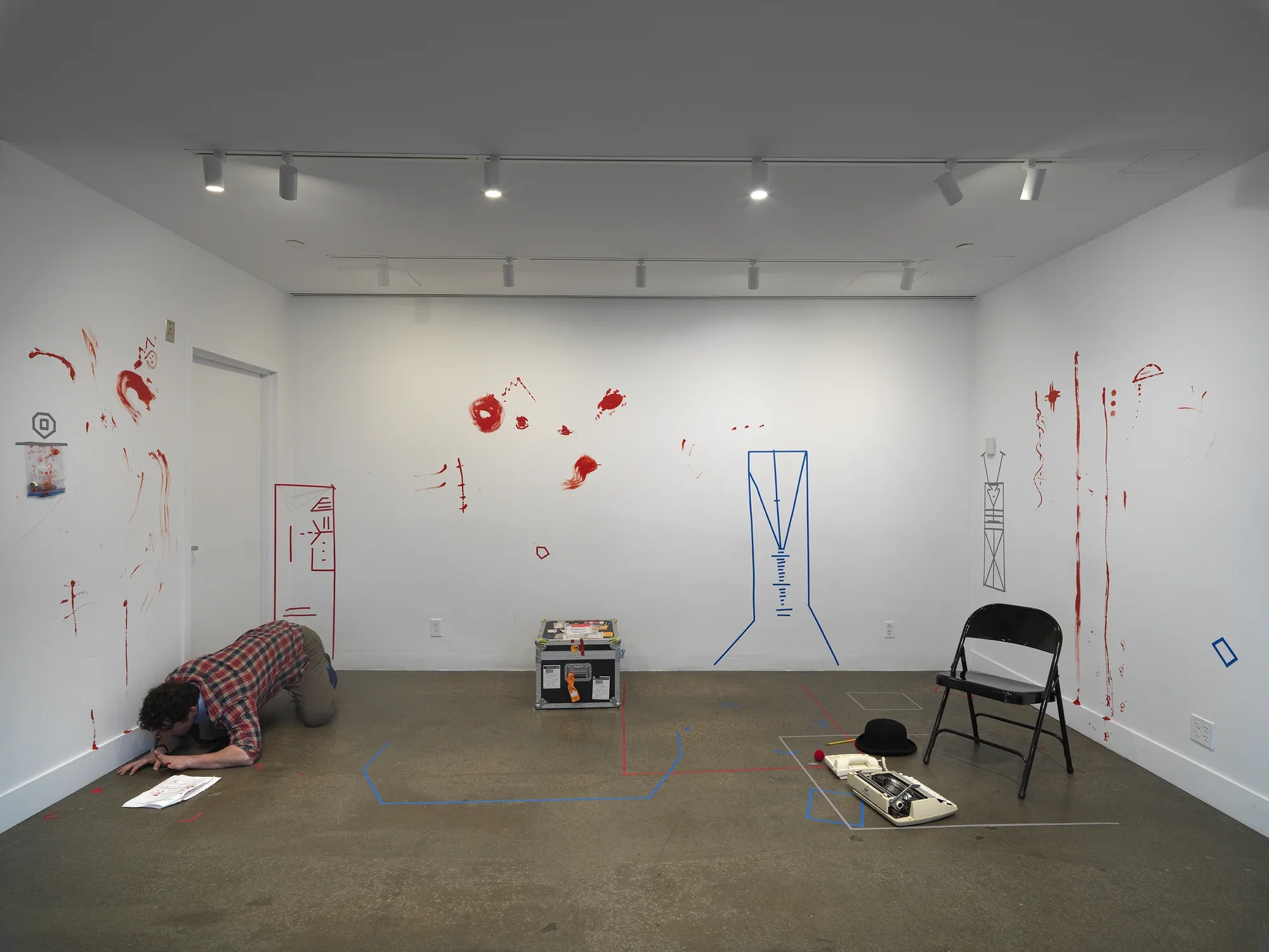A man in a plaid shirt, resting on his arms and knees, occupies the corner of a gallery. A printed script sits on the floor in front of him. The floor has some geometric lengths of colored tape and a few props for the performance, including folding chair, a typewriter, a top hat, a clown nose, and a traveling case.