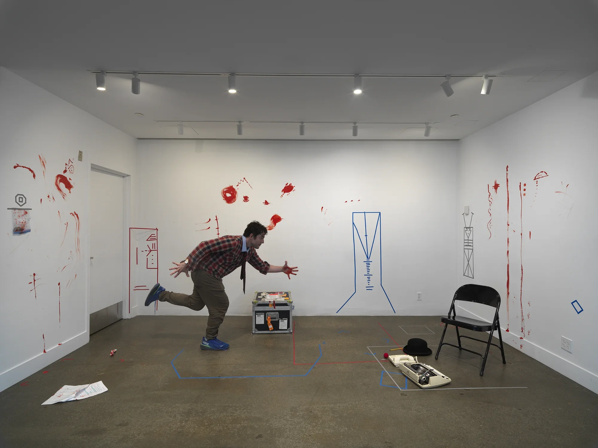 A man in a shirt and tie, viewed in profile, travels exaggeratedly across a gallery space: his left leg is lifted behind him, perhaps moving quickly, perhaps frozen in time. The palm and splayed fingers of one of his hands is red with paint. He is surrounded by the props and traces of the performance, including tape and paint on the walls.