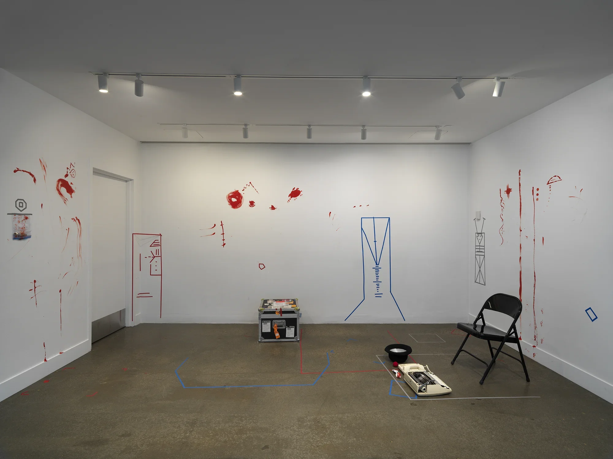A view of an installation—perhaps a spare theatrical set—in a white-walled gallery. The concrete floor space is organized by overlapping lengths of colored tape; a metal folding chair, a typewriter, a top hat, and a trunk covered in shipping labels sit on the floor. The walls are marked by obscure geometric figures—resembling buildings or machines—made of more colored tape and other expressive smears and trails of red paint.