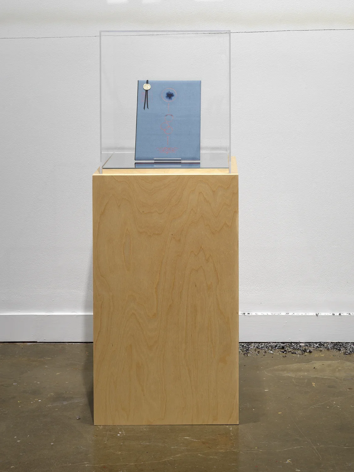 A blue volume—its cover marked by a wax seal and an abstract design made of a few circles and lines—is encased in glass atop a wooden pedestal in a gallery.
